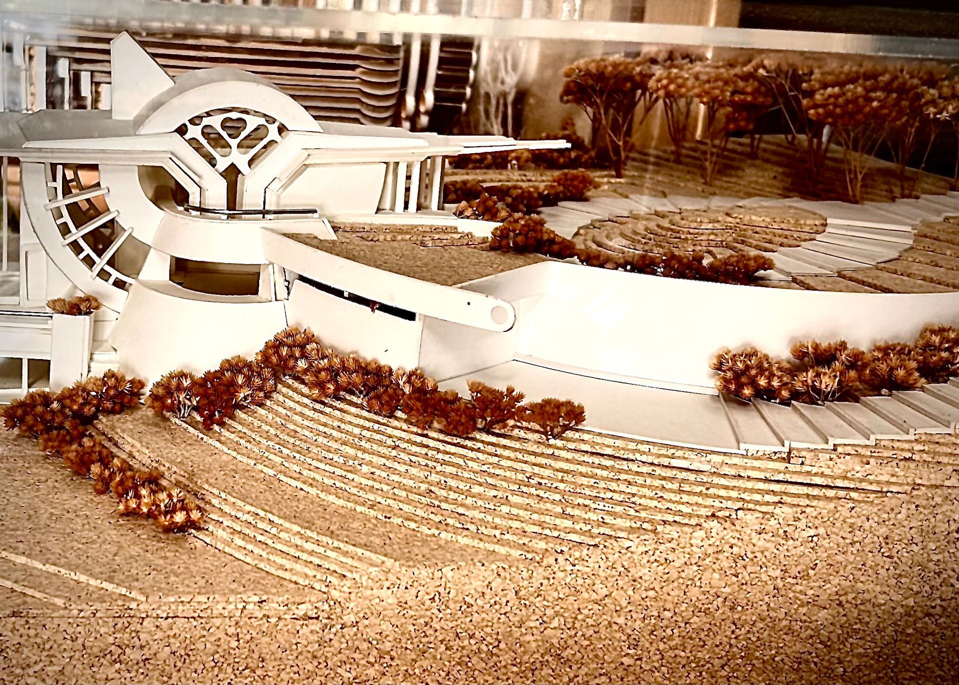 Curved white modern building model with terraced landscaping and small trees in a plaza.