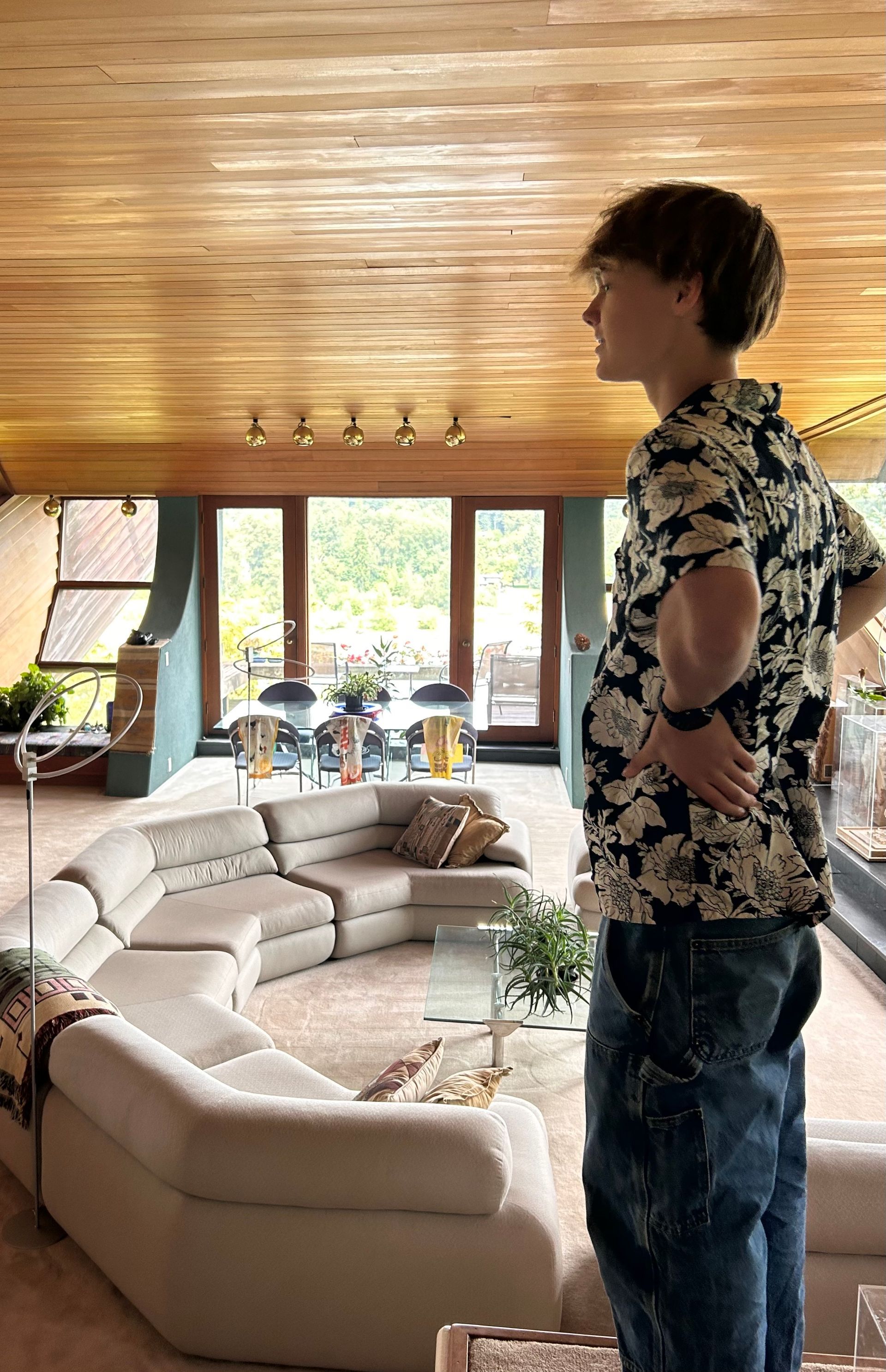 Person standing in a bright living room with large windows, beige sectional sofa, and wood-paneled ceiling