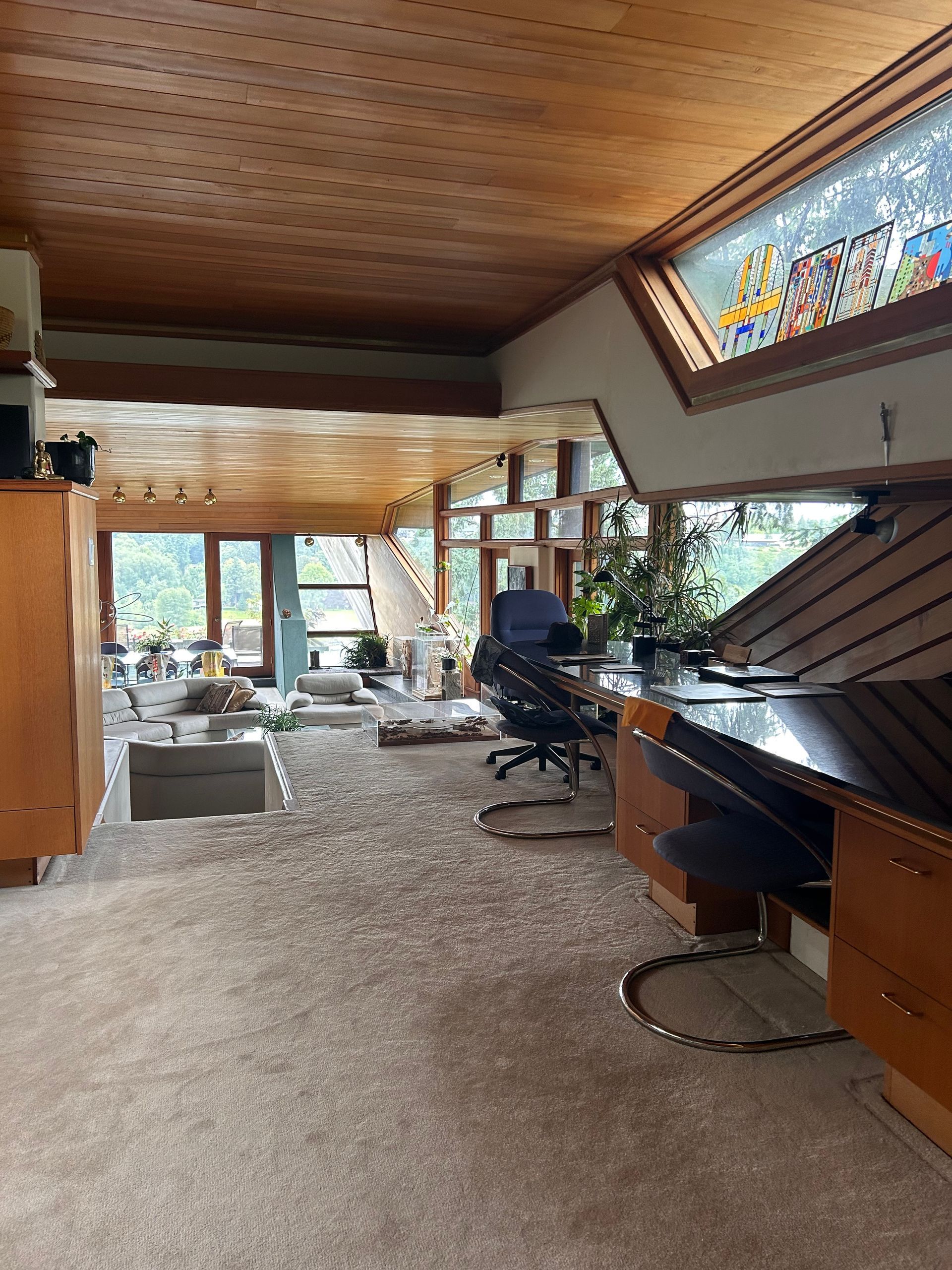 Spacious sunlit living room with wood ceiling, large windows, carpet, and a piano near a desk.
