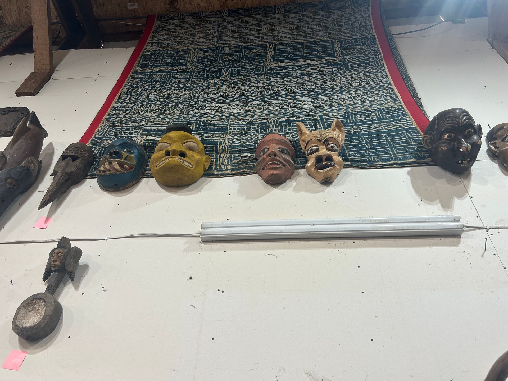 Six colorful carved masks and small tools arranged on a white floor beside a patterned rug.