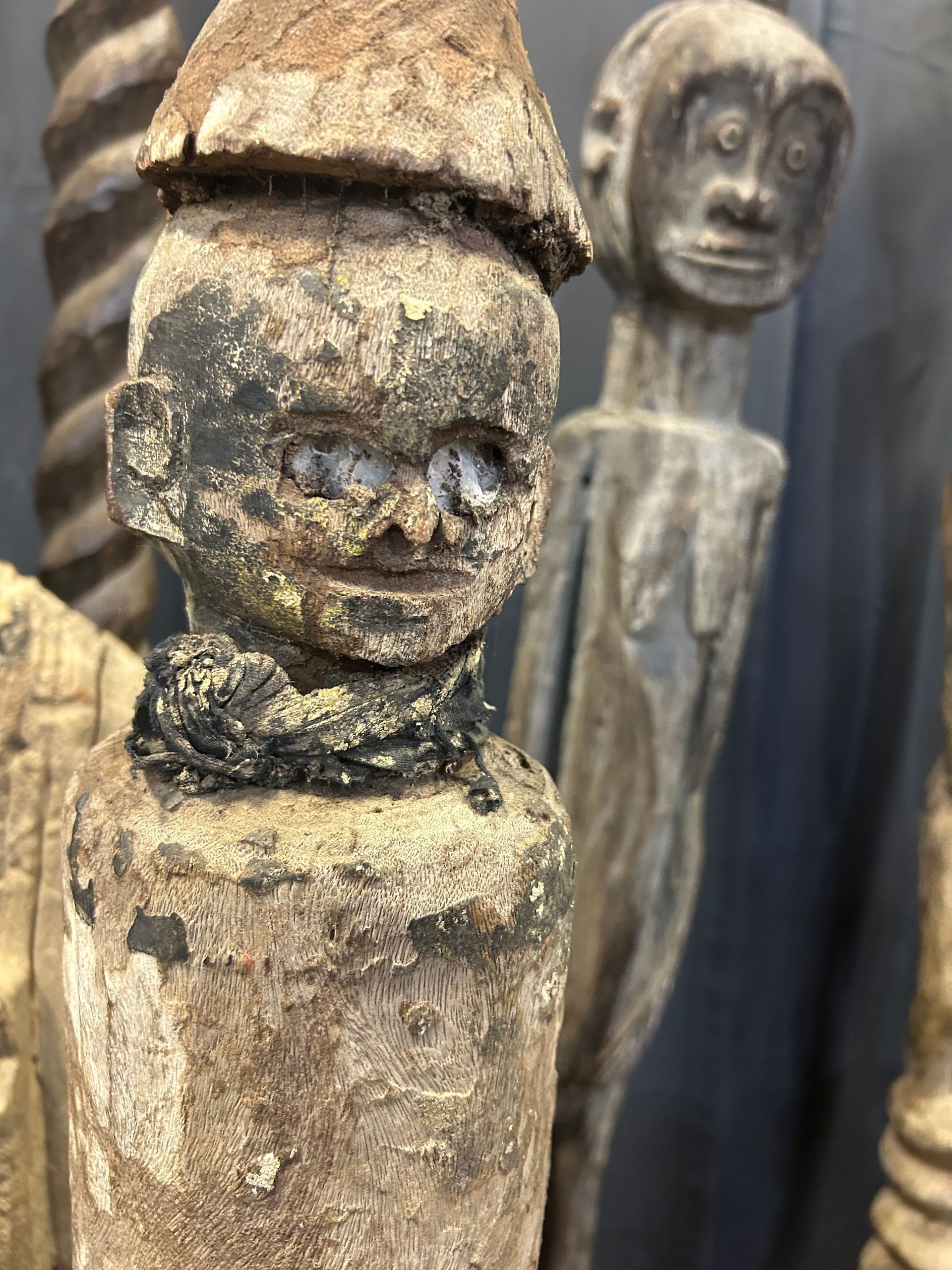 Close-up of weathered wooden carved figures, one in front with a conical hat and blurred figures behind