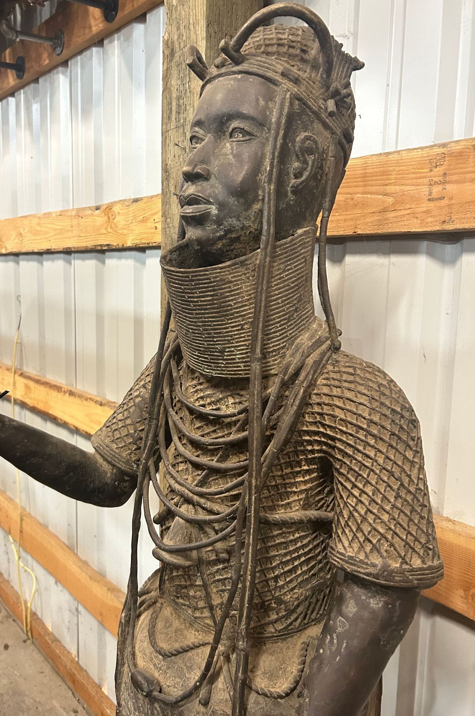 Bronze statue of a seated figure with intricate jewelry and headdress indoors against a metal wall