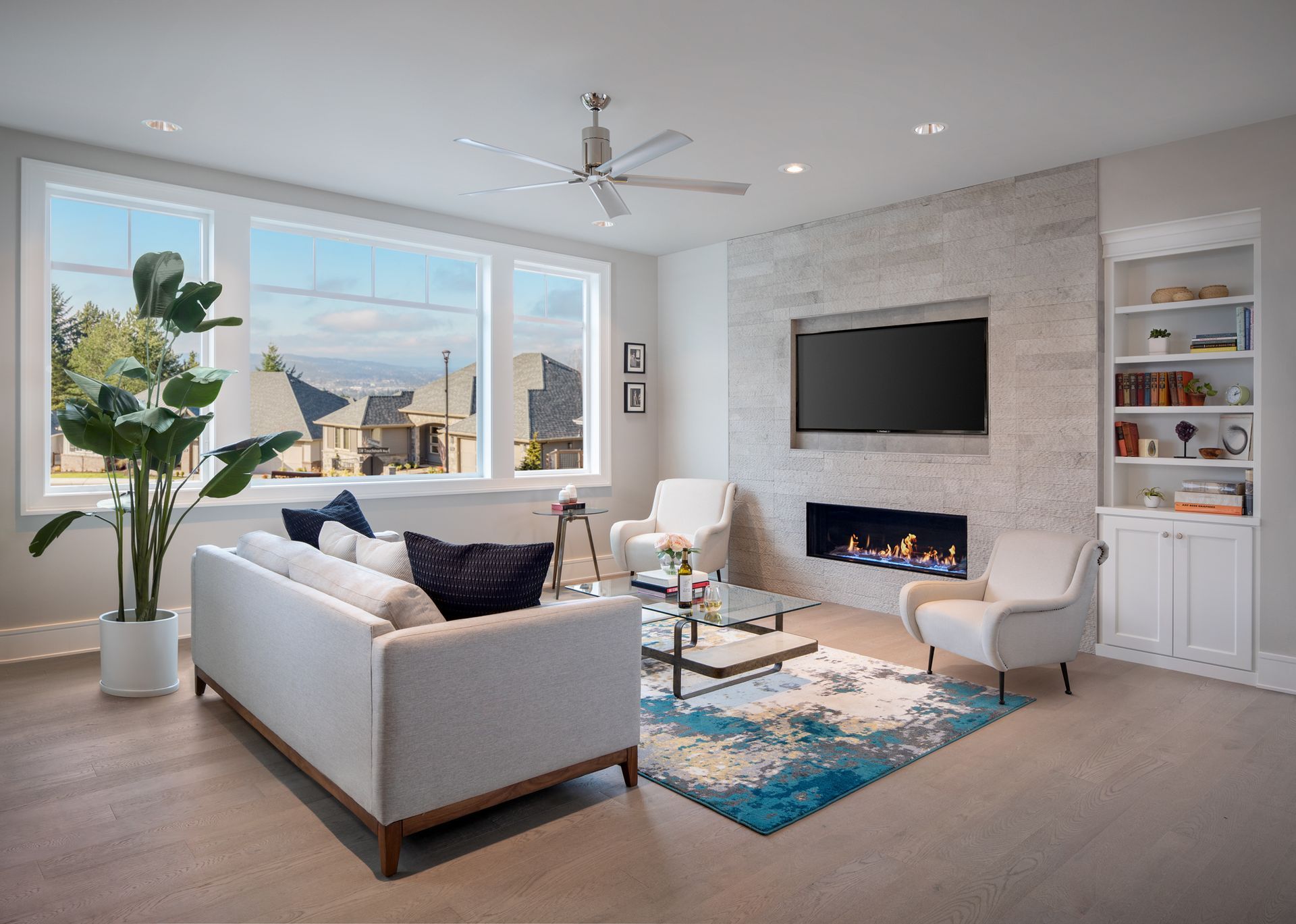 Bright modern living room with large windows, white sofa, wall TV, fireplace, and blue patterned rug