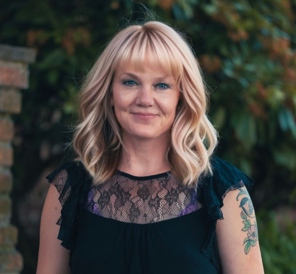Smiling woman in a black lace top with blonde hair, standing outdoors against a blurred leafy background