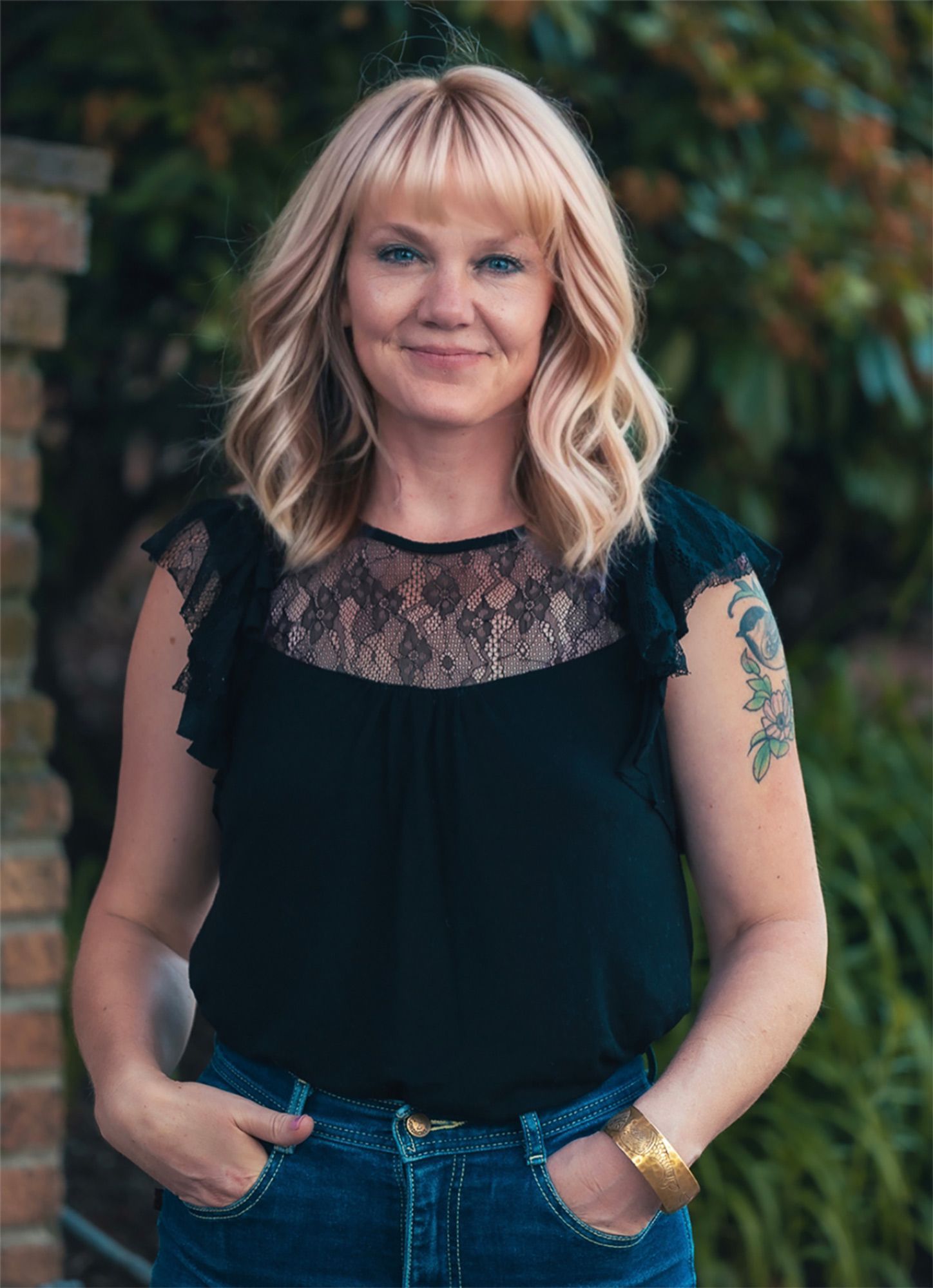 Woman standing outdoors with hands in jeans pockets, wearing a black lace top and smiling.