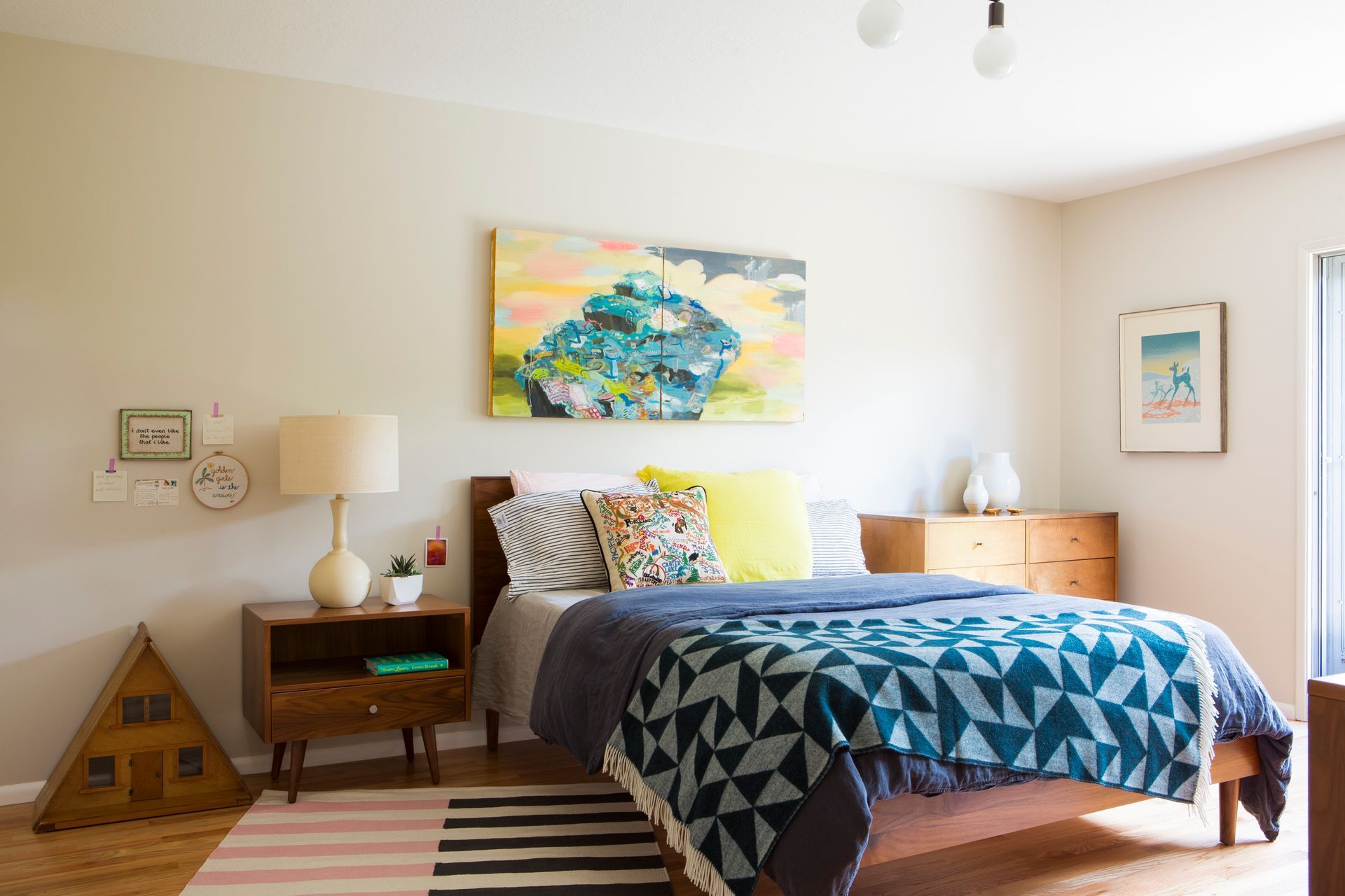 Bright bedroom with a bed, colorful wall art, wooden nightstand, and striped rug