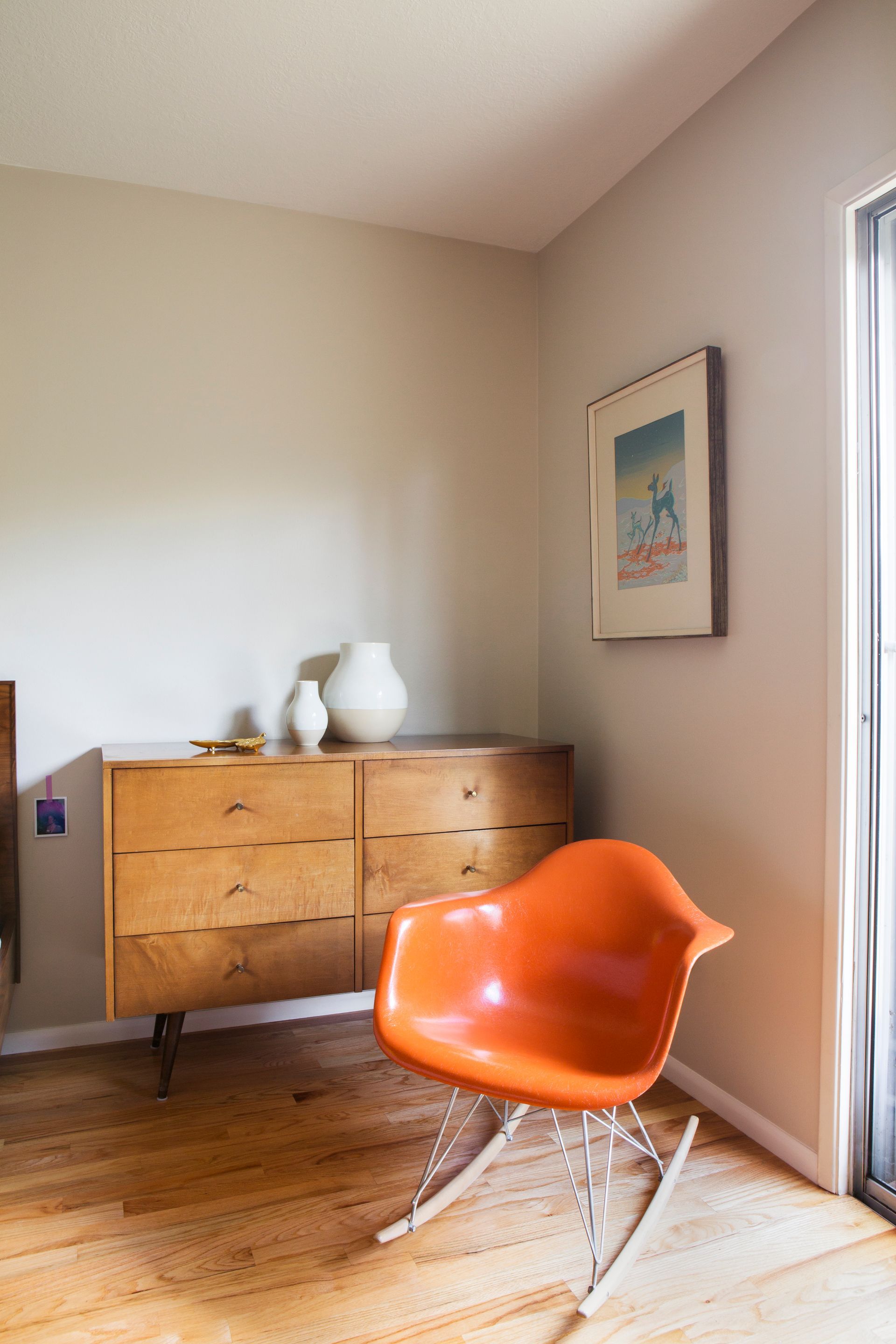 Minimalist room with orange chair, wooden dresser, and framed wall art by a sliding glass door