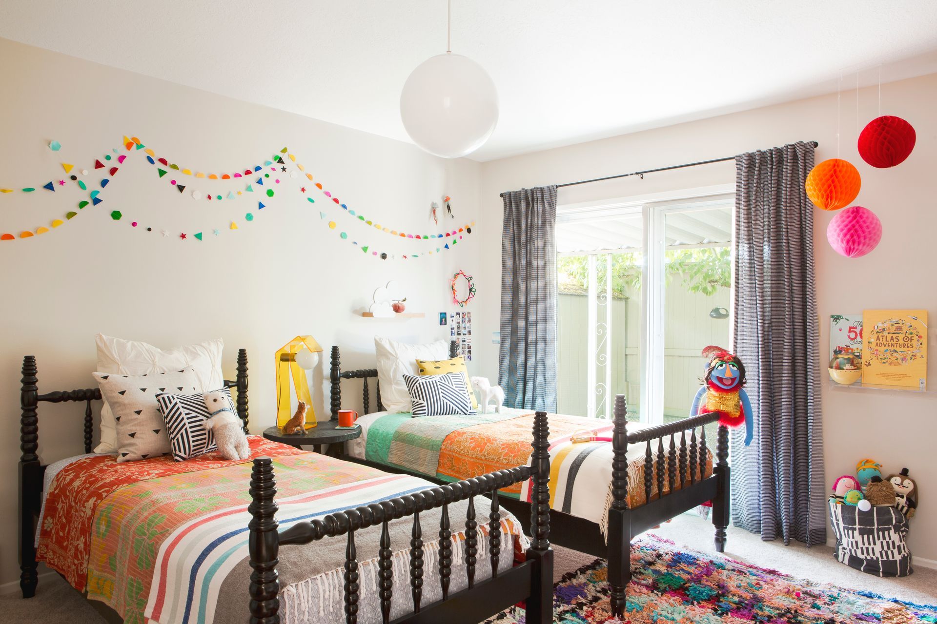 Colorful twin bedroom with bunting, pom-poms, patterned rugs, and a window with curtains
