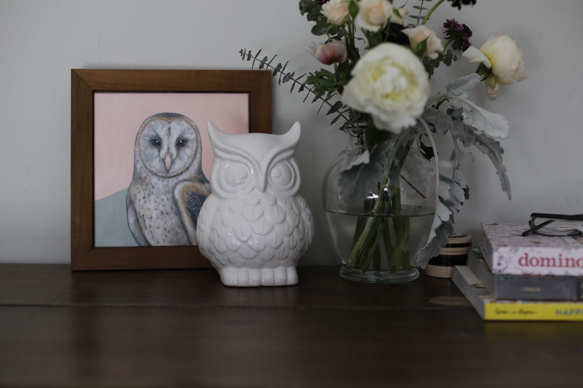 Framed owl illustration, white owl figurine, and vase of flowers on a dark shelf beside stacked books