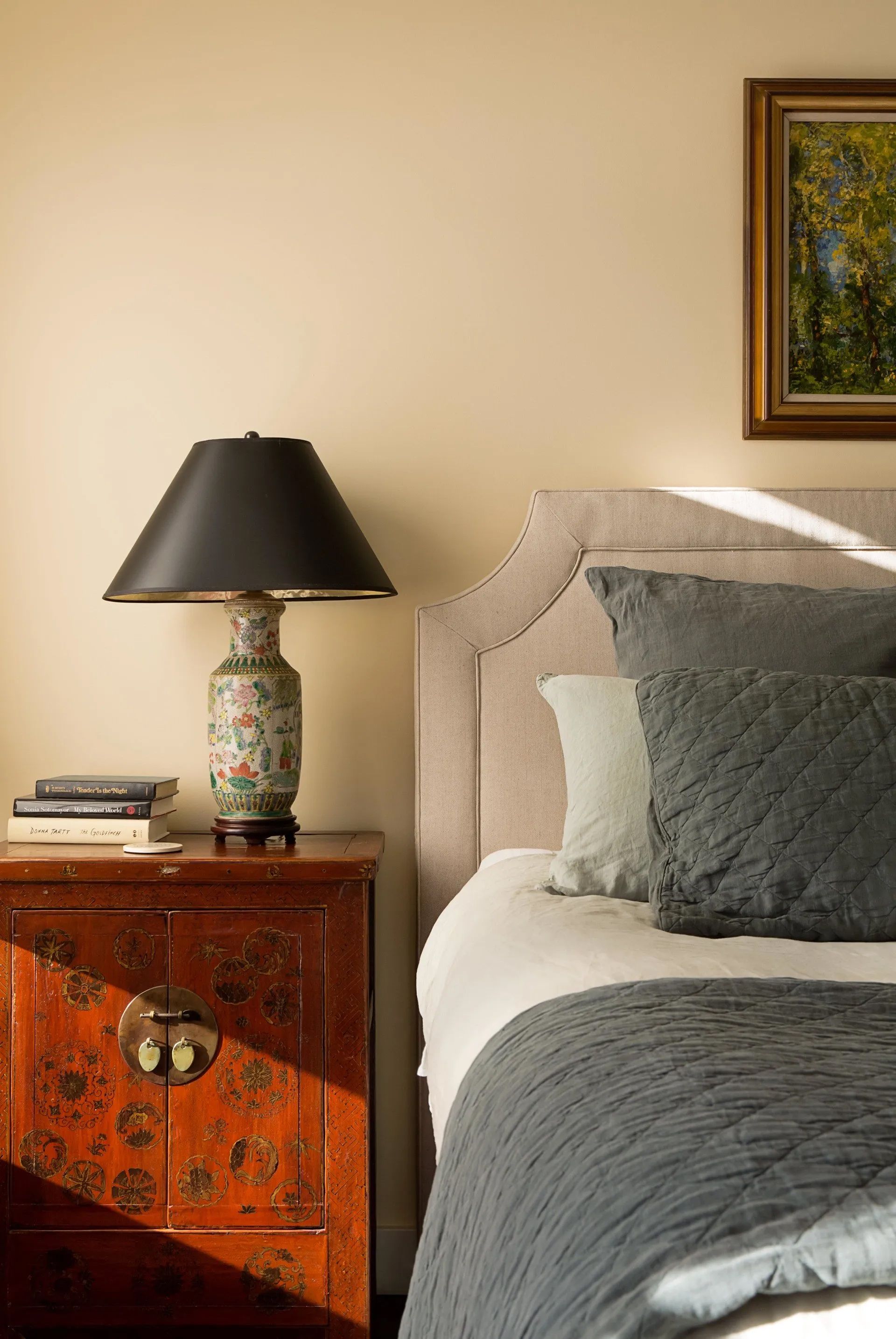 Bedroom corner with beige upholstered bed, patterned nightstand, black lamp, and framed wall art.