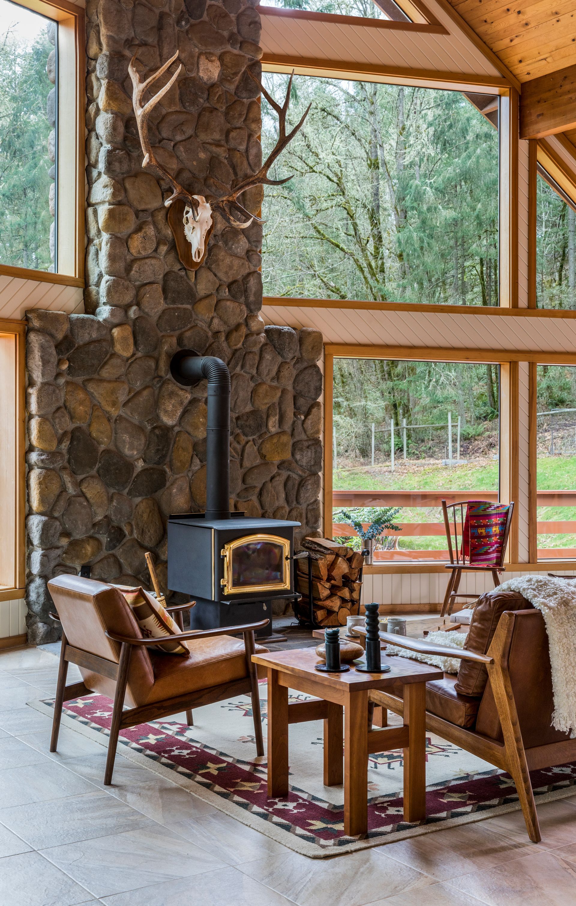 Rustic cabin living room with stone fireplace, large windows, and wooden chairs by a red rug.