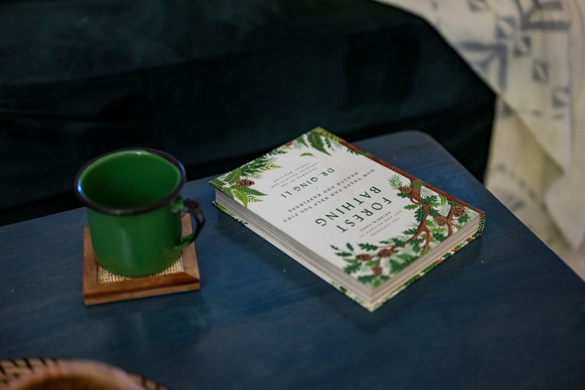 Green mug on a coaster beside a floral-covered book on a blue table.
