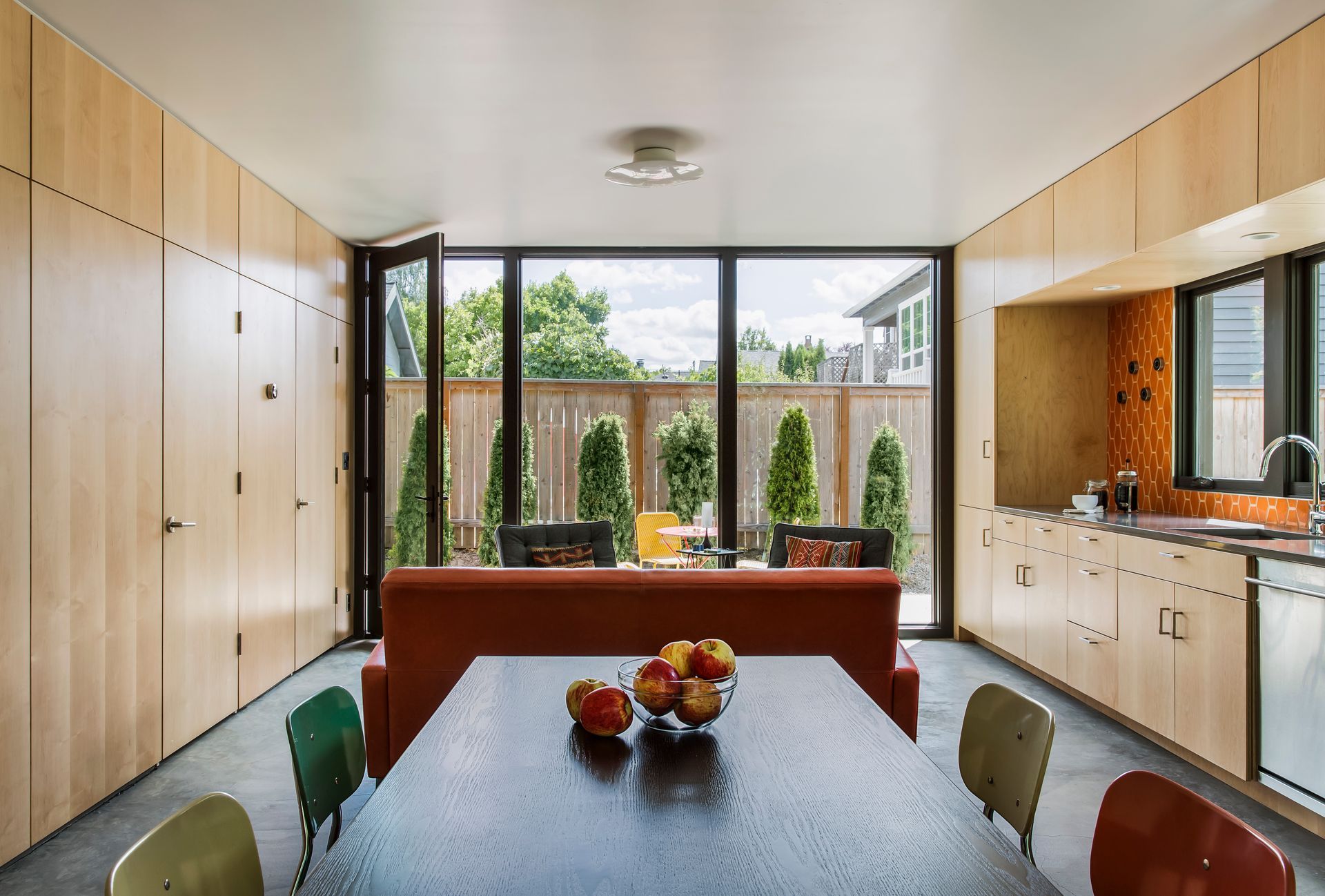 Modern kitchen with wooden cabinets, central dining table, and large windows overlooking a garden.