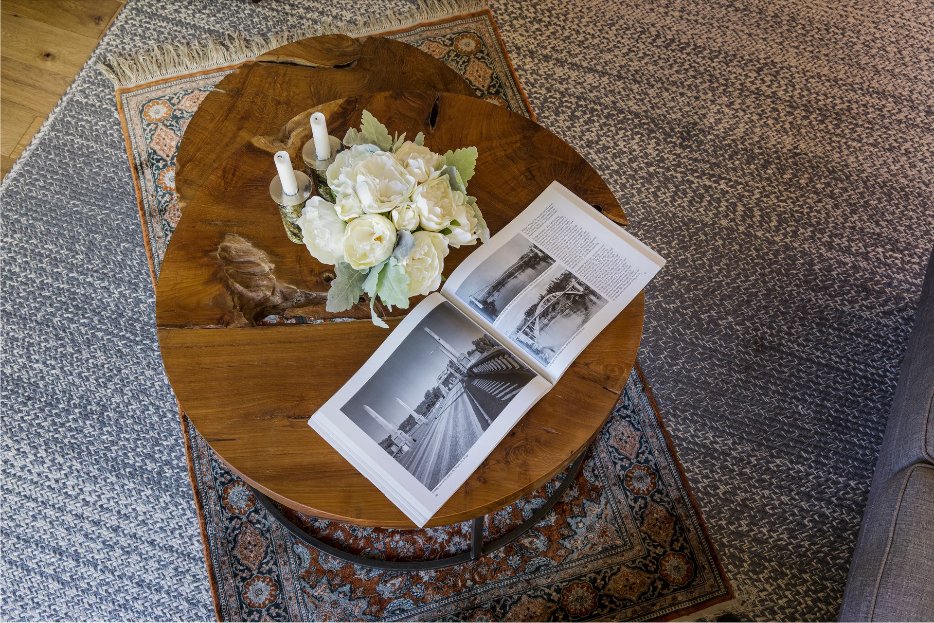Round wooden coffee table with white flowers and a black-and-white photo booklet on a patterned rug.