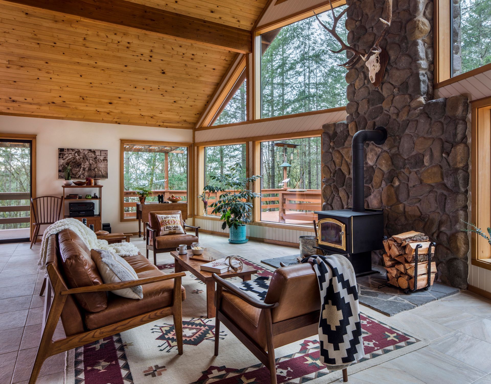Cozy rustic living room with wood ceiling, stone fireplace, and large windows overlooking trees