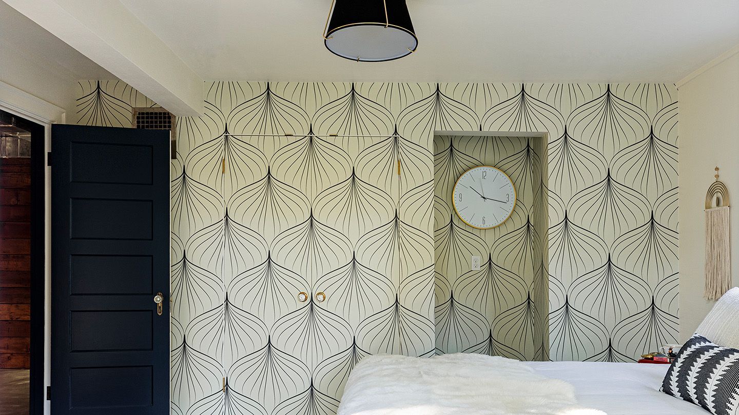 Bedroom with patterned wallpaper, dark door, round clock, and a white bed with striped pillows.