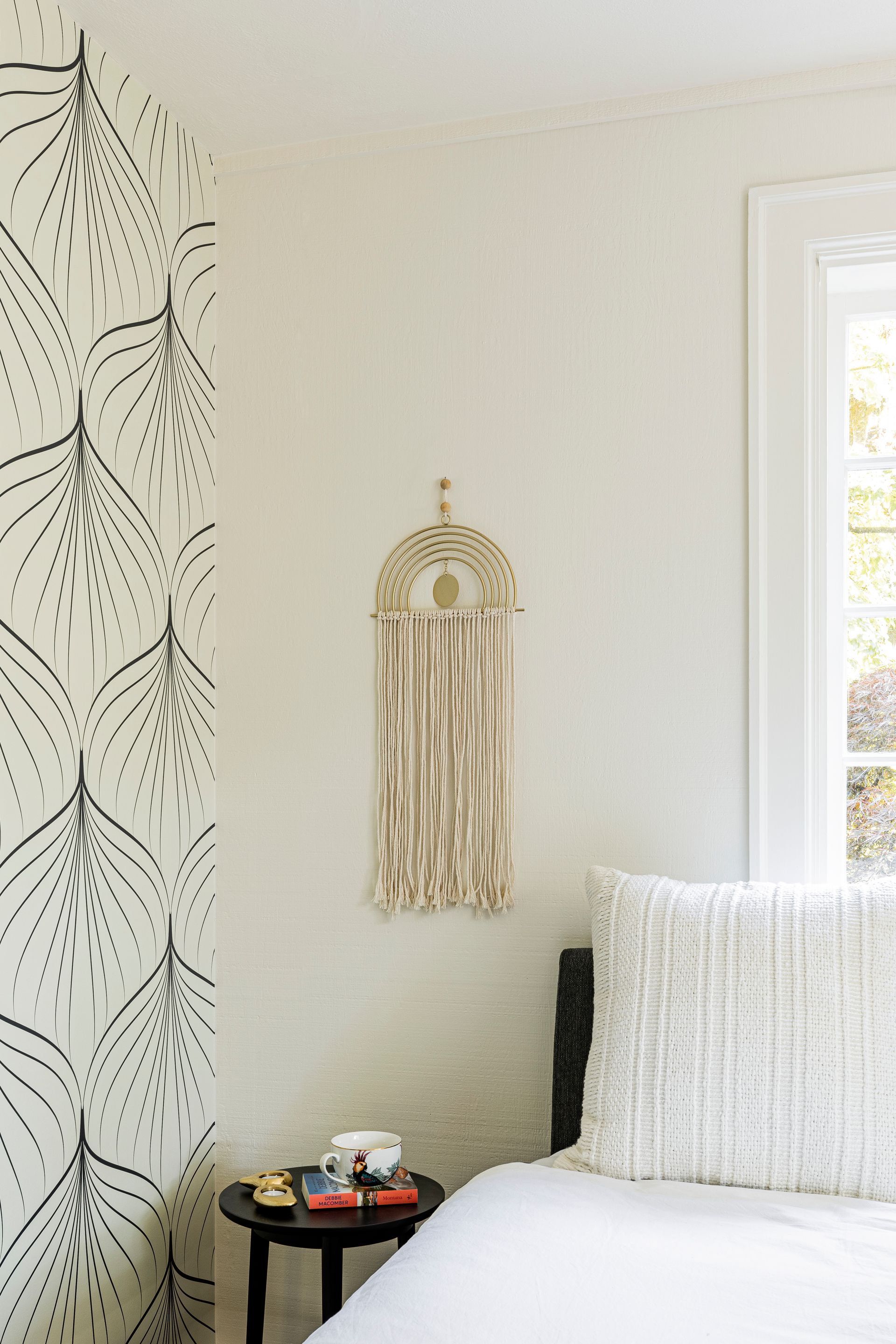 Cream bedroom corner with macramé wall hanging, black side table, and bed by a window.