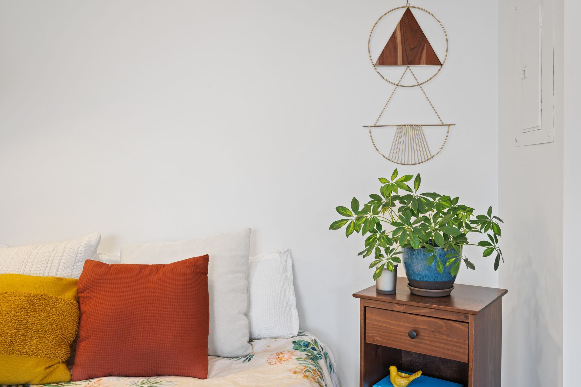 Cozy white bedroom corner with colorful pillows, a potted plant on a nightstand, and hanging wall decor