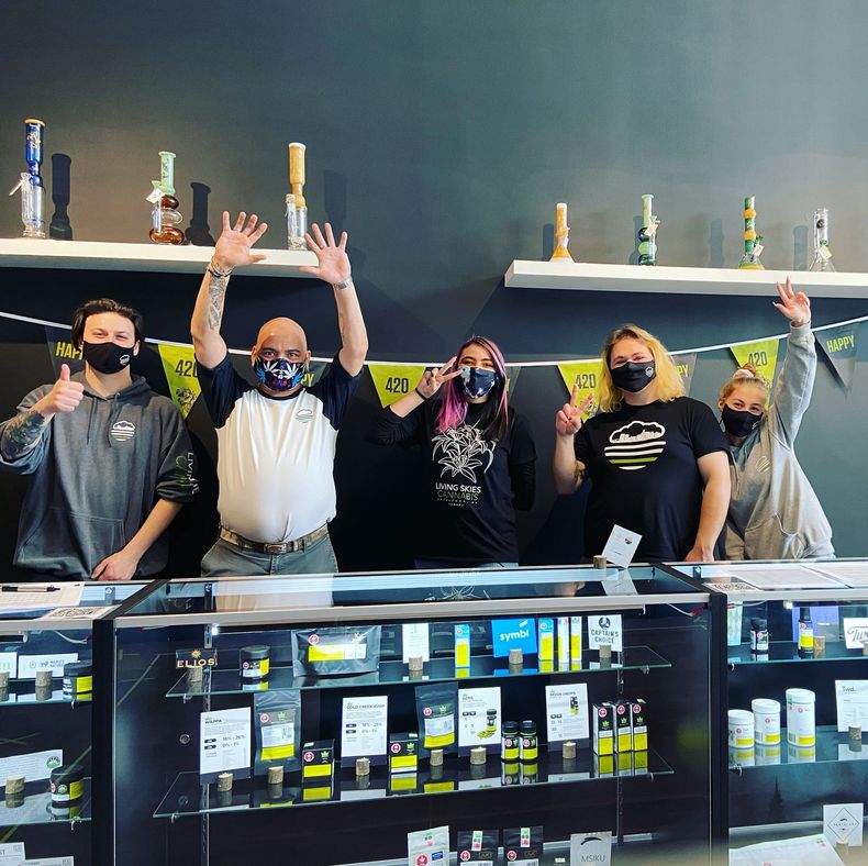 Five masked people posing behind a glass display case, some with arms raised, in a shop interior.