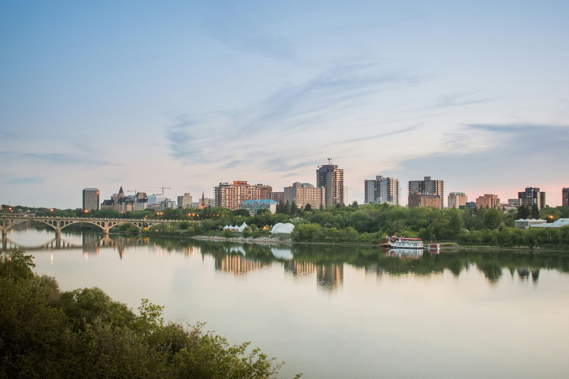 City skyline and bridge over a calm river at sunset, with trees in the foreground.
