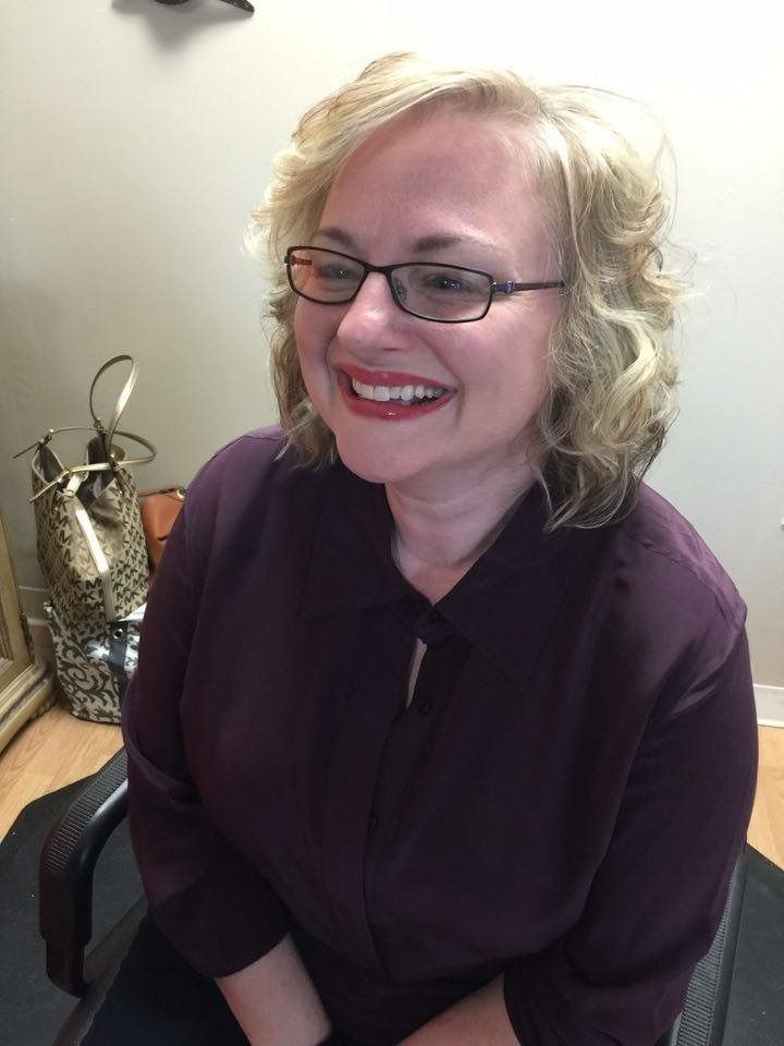 Smiling woman with glasses seated indoors, wearing a dark purple blouse.