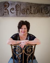Woman smiling behind ornate chair beneath a sign reading “Reflections”