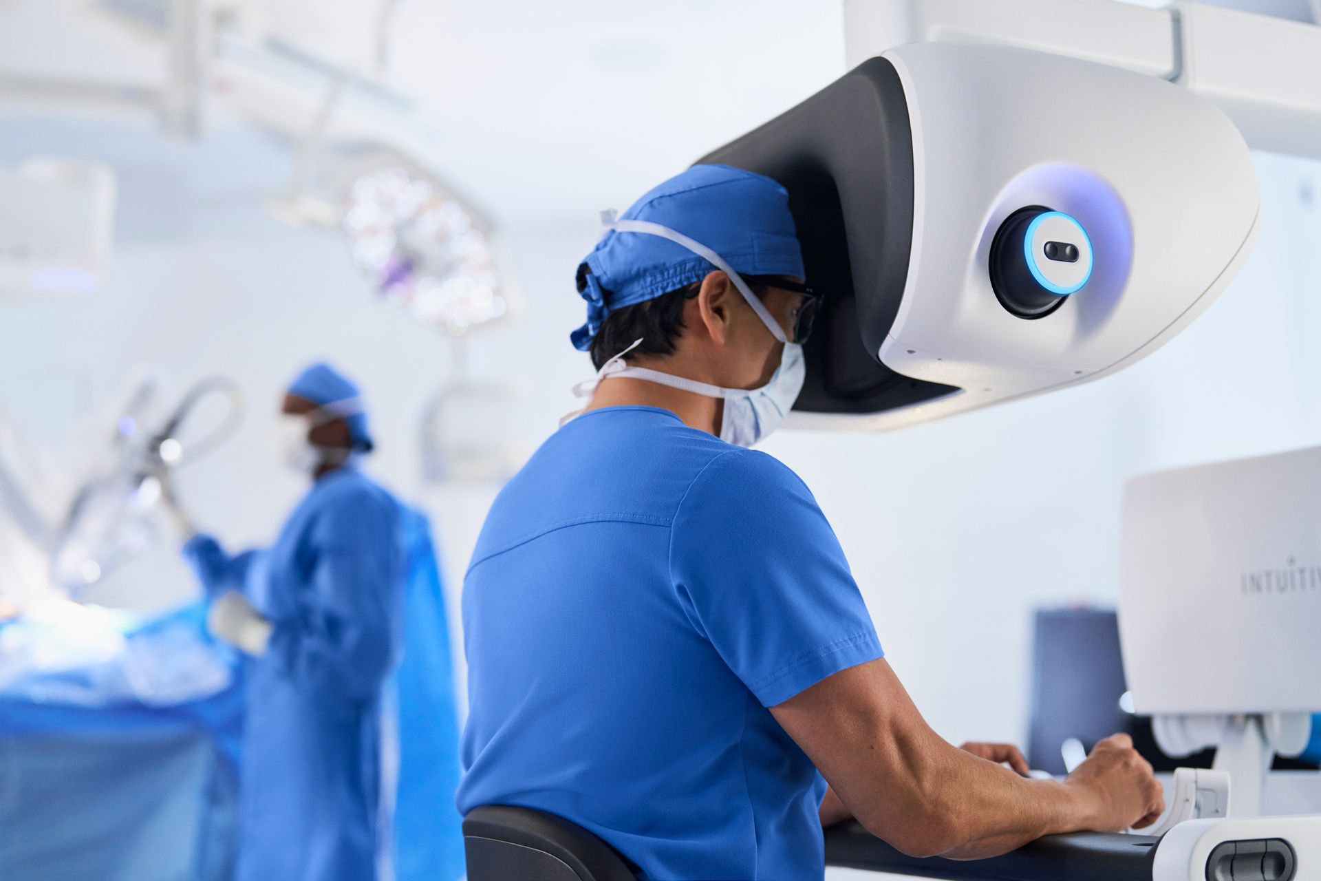 Cirurgiões de jaleco azul operando com equipamento de cirurgia robótica em uma sala de cirurgia bem iluminada.