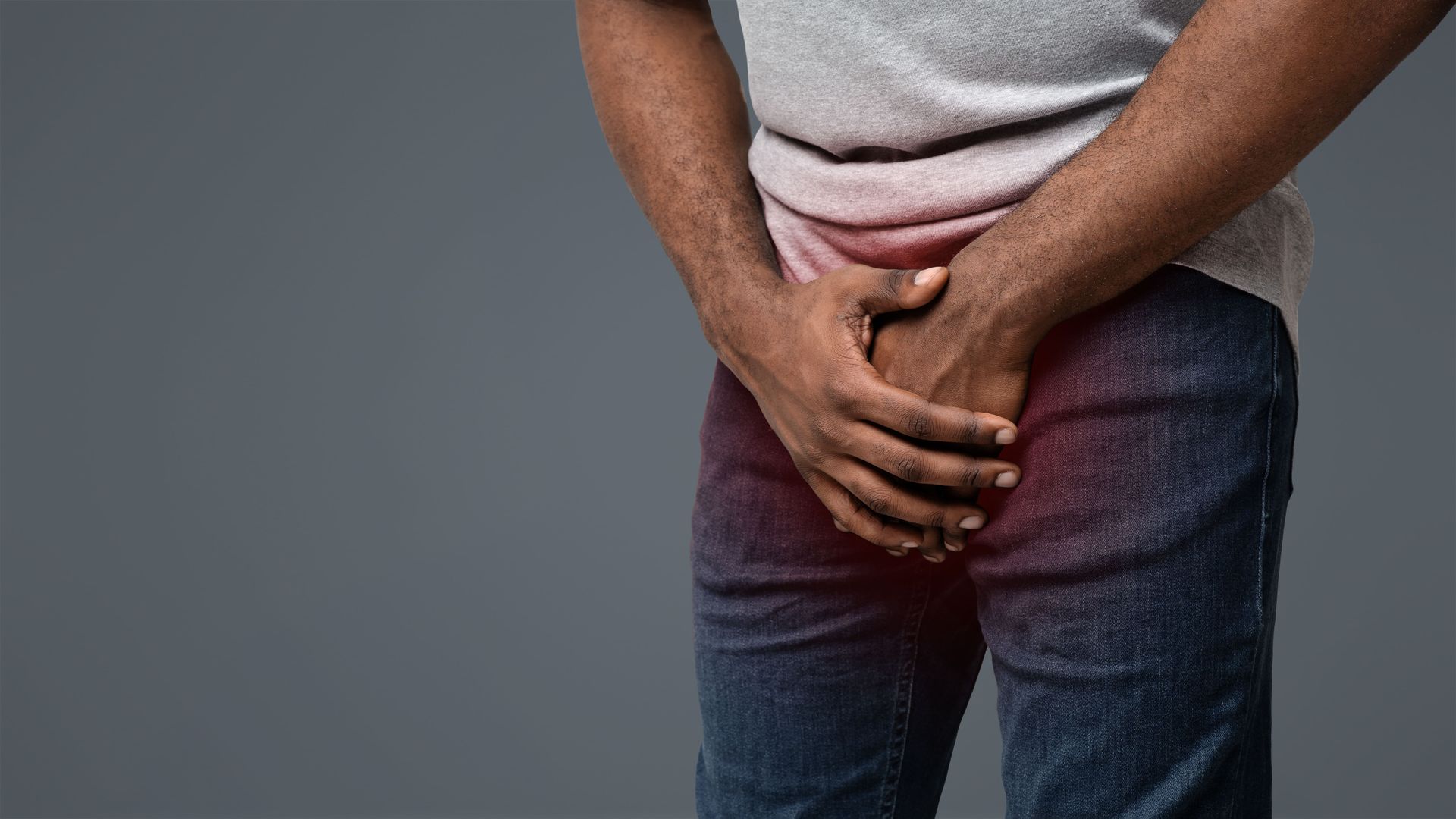 Pessoa em pé com as mãos cruzadas sobre a virilha, vestindo uma camisa cinza e calça jeans escura, em um fundo cinza.