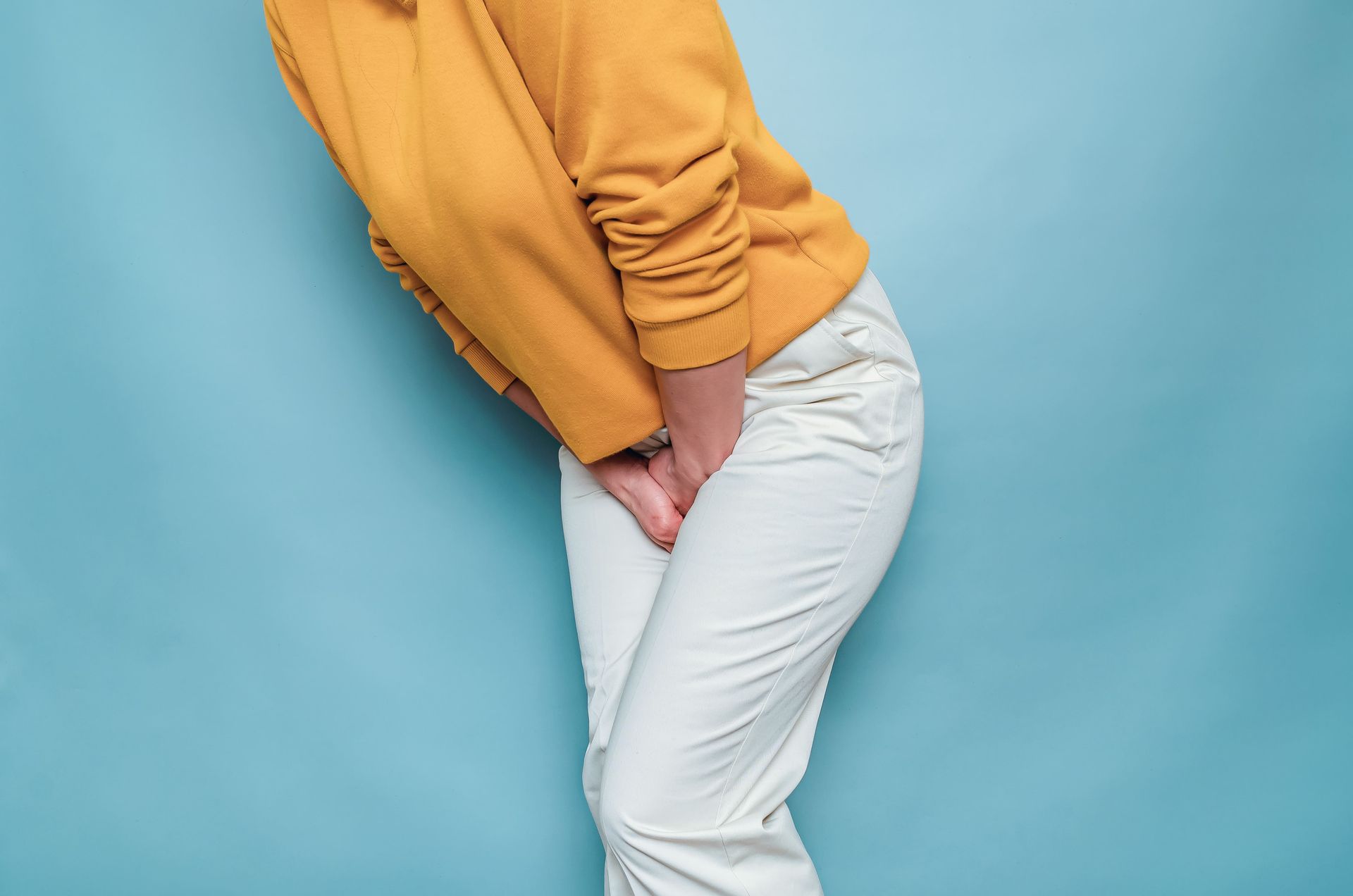 Pessoa vestindo suéter mostarda e calça branca, inclinada para a frente contra um fundo azul.