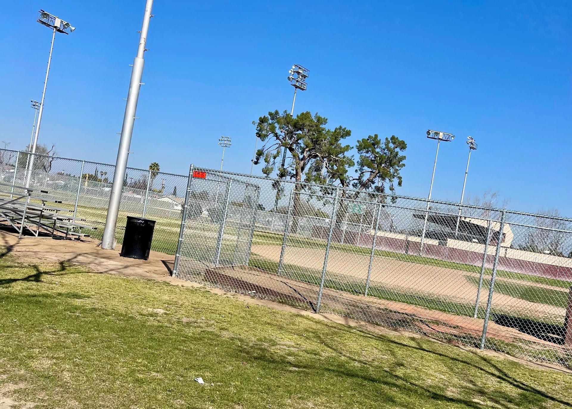 Chain-link fence around a baseball field under a bright blue sky, with light poles and a few trees.