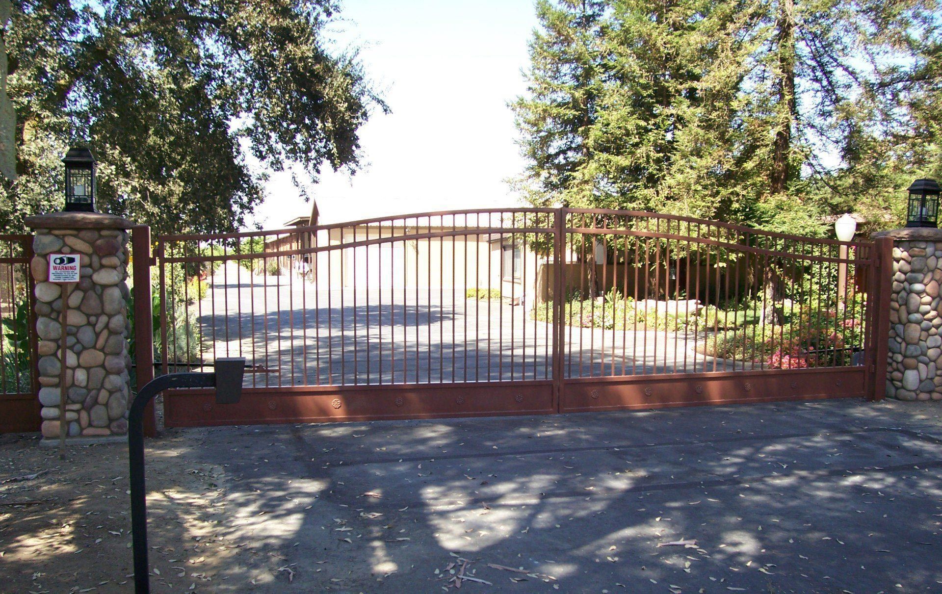 Closed iron driveway gate between stone pillars, with trees and bright sunlight behind it.