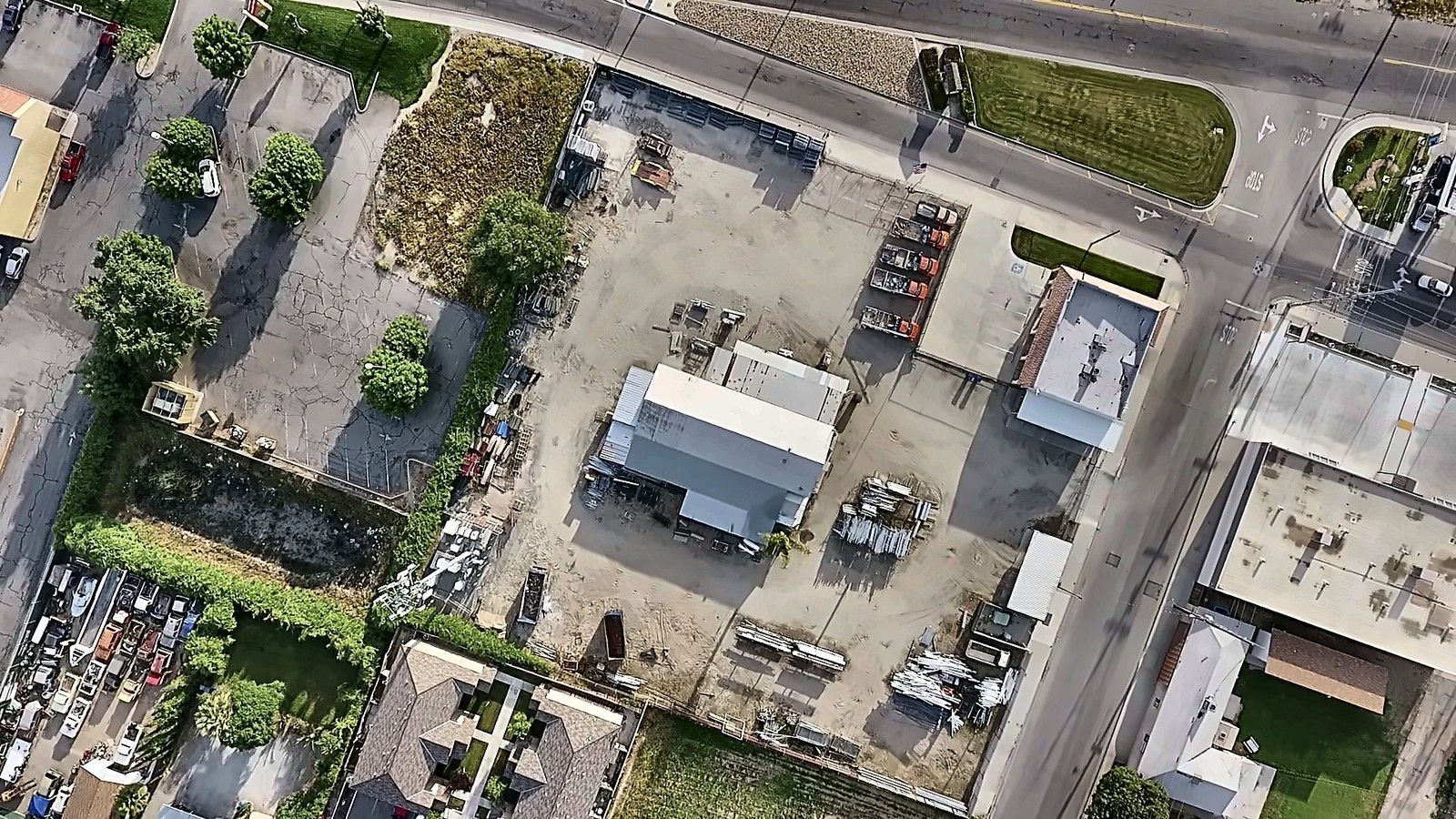 Aerial view of a residential lot with a small house, parked vehicles, trees, and surrounding homes.