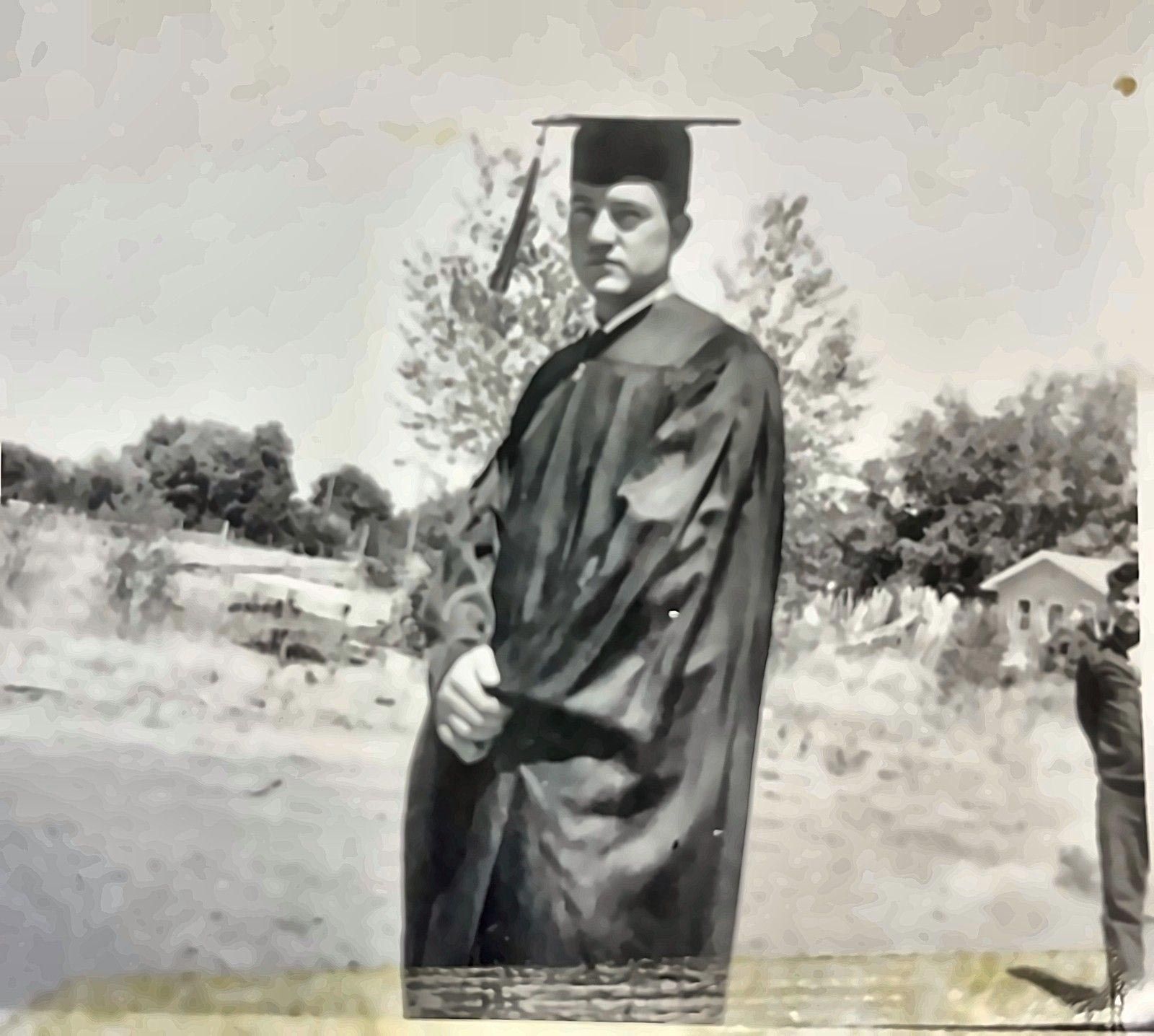 Person in a black graduation gown and cap standing outdoors in a grassy area.