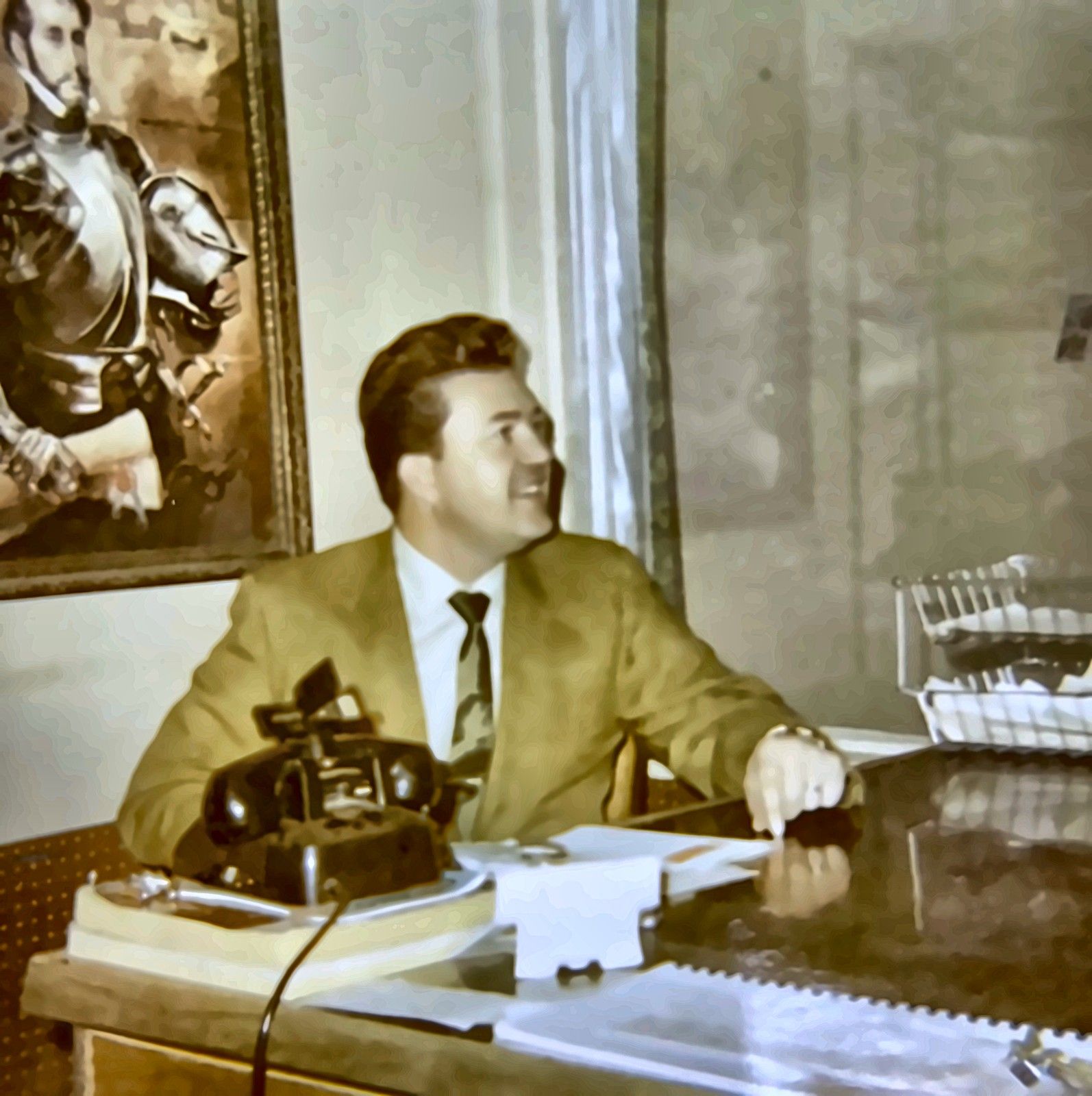 Man in a tan suit sitting at a desk, looking left beside a black rotary phone and papers
