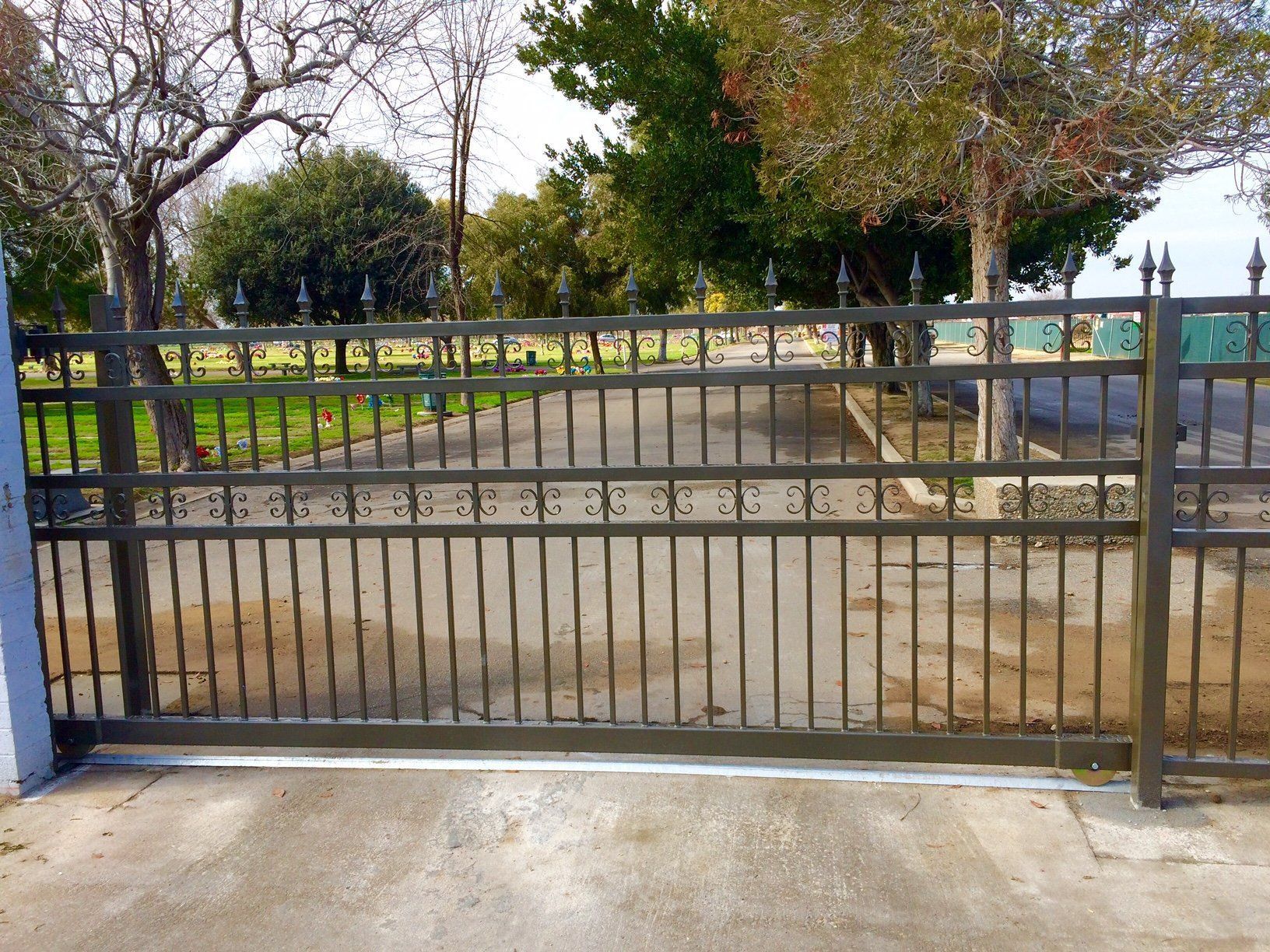 Black metal fence along a driveway beside a house with stone walls and red-tiled roofs