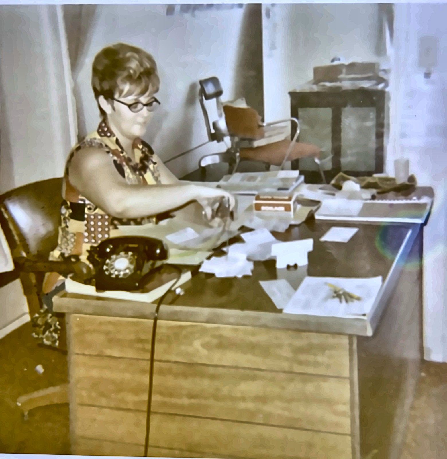 Woman seated at a desk using a rotary phone, surrounded by papers and office items.