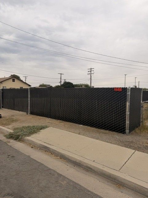 Chain-link fence with black privacy screen along a sidewalk under a cloudy sky.