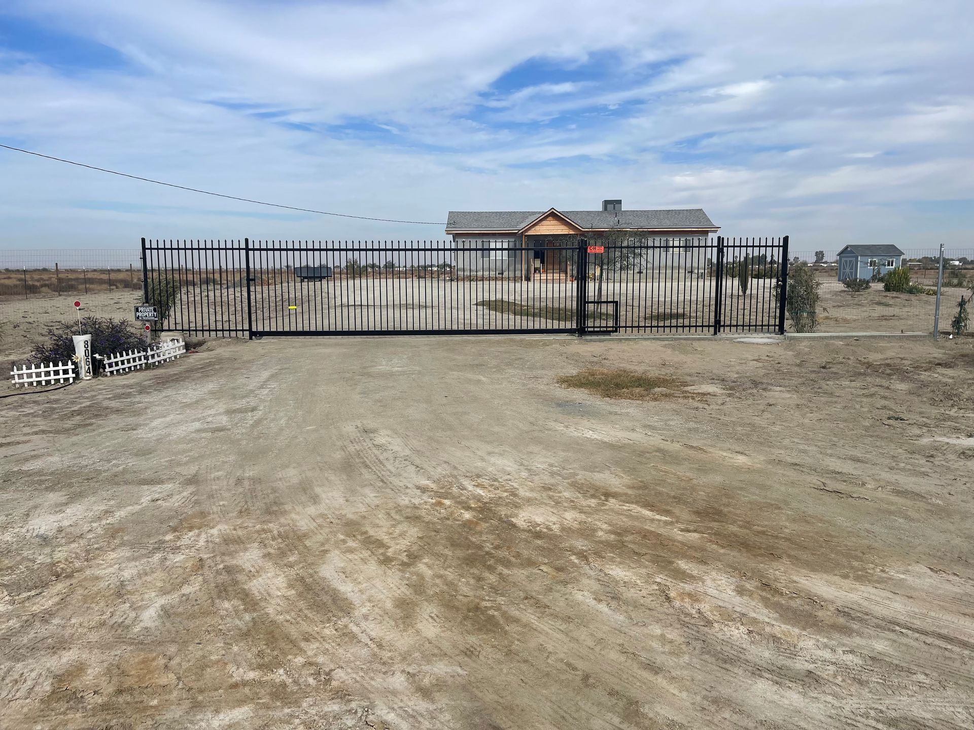 Small white house behind a black fence on a barren dirt lot under a cloudy sky