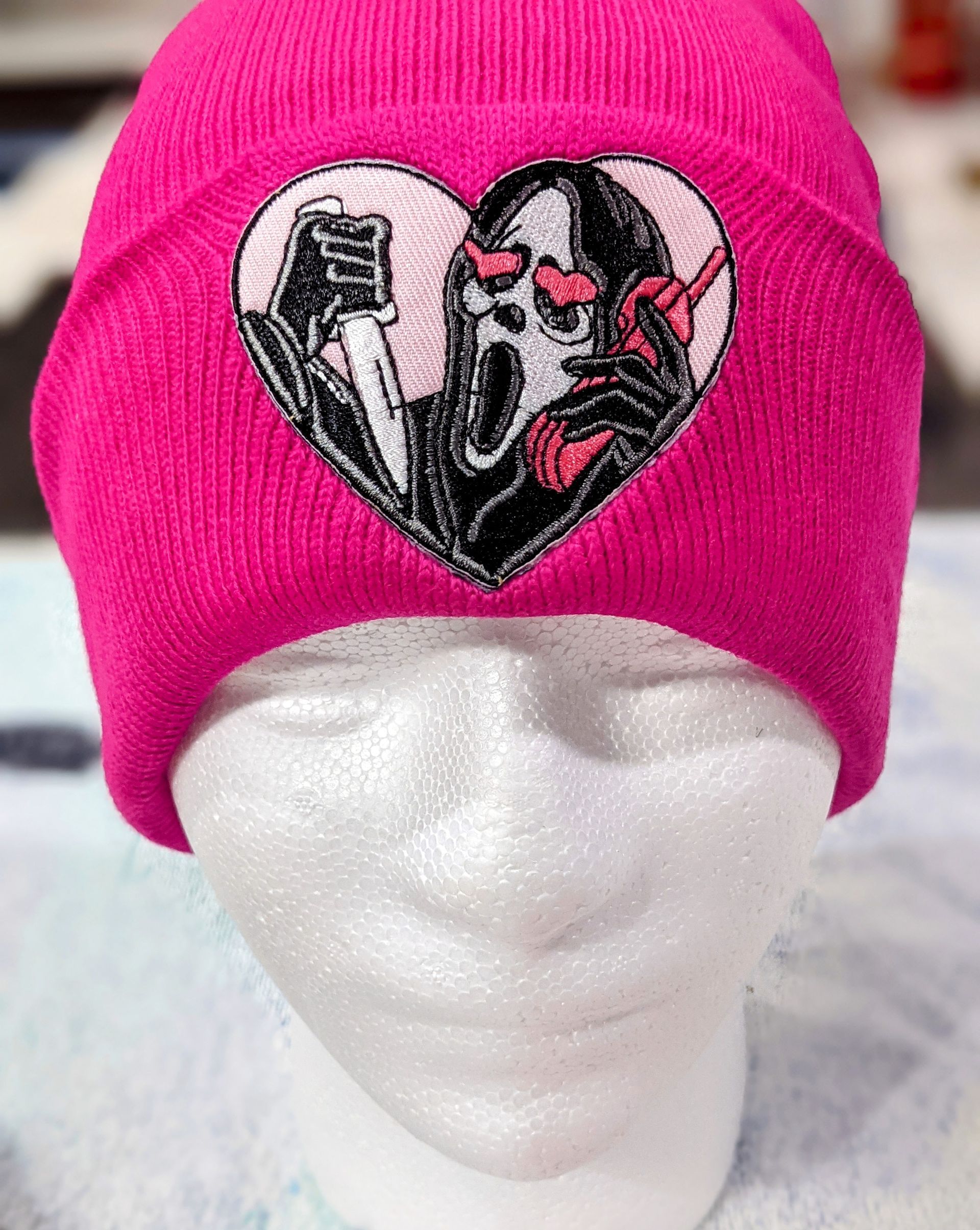 A bright pink beanie featuring an embroidered patch of the Ghostface mask with heart-shaped eyes, holding a knife and phone.