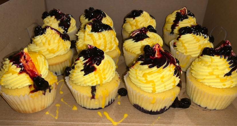 Yellow cupcakes with yellow frosting and dark cherry topping in a cardboard box.