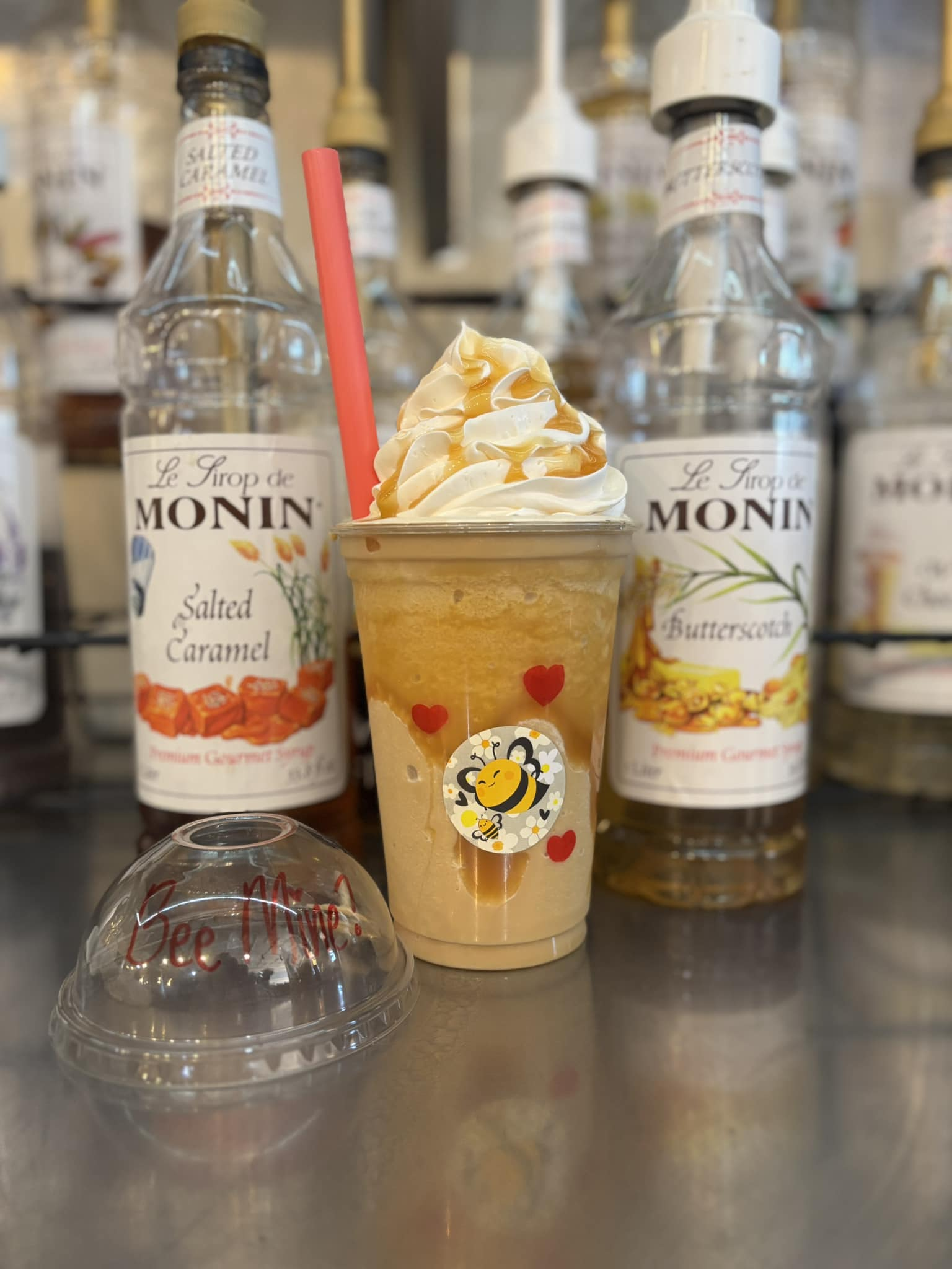 Frosted drink with whipped cream, caramel drizzle, straw, and bee sticker; bottles of syrup in background.