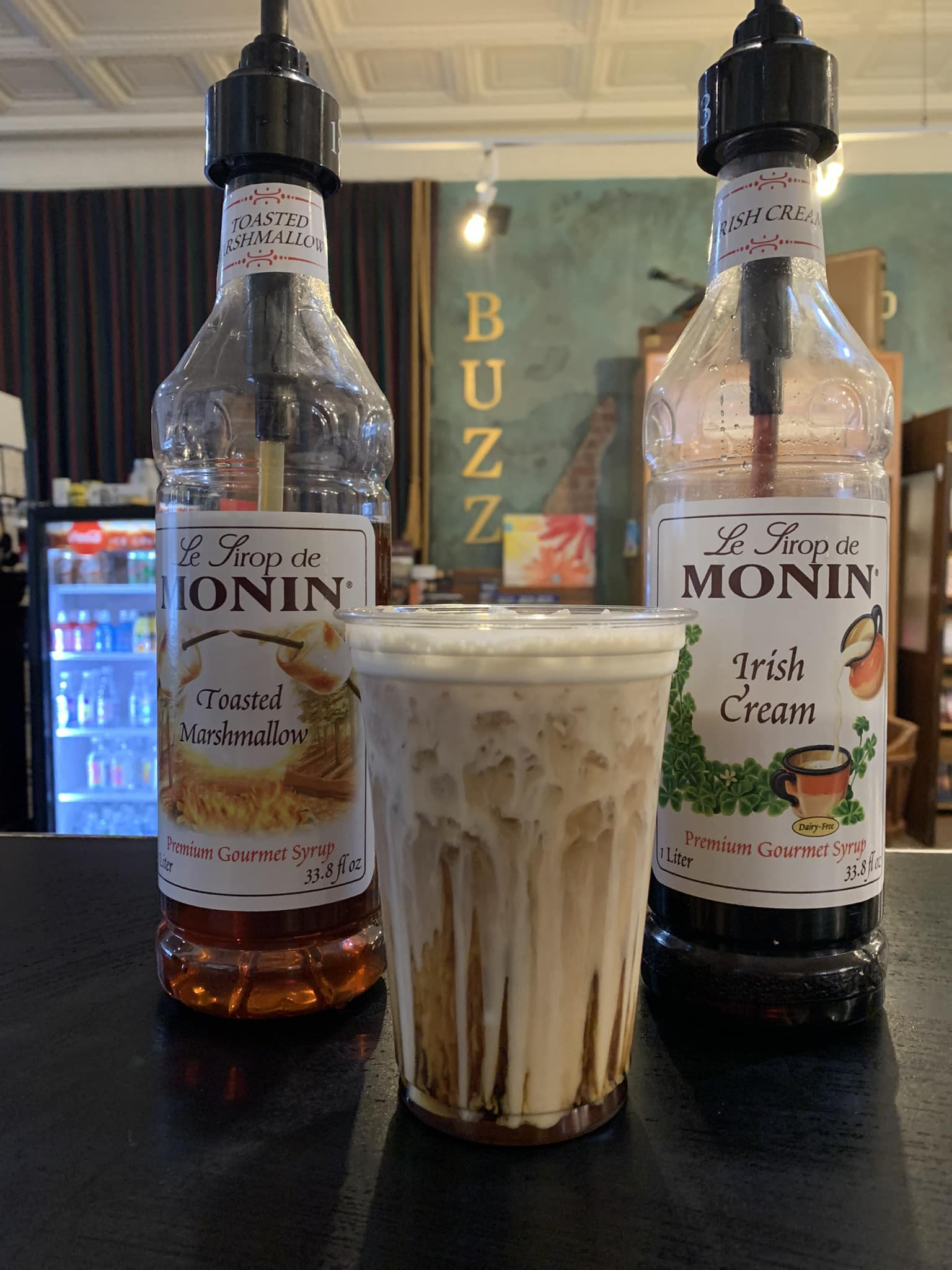 A cup of iced coffee sits between two bottles of Monin syrups, caramel and Irish cream, on a bar.