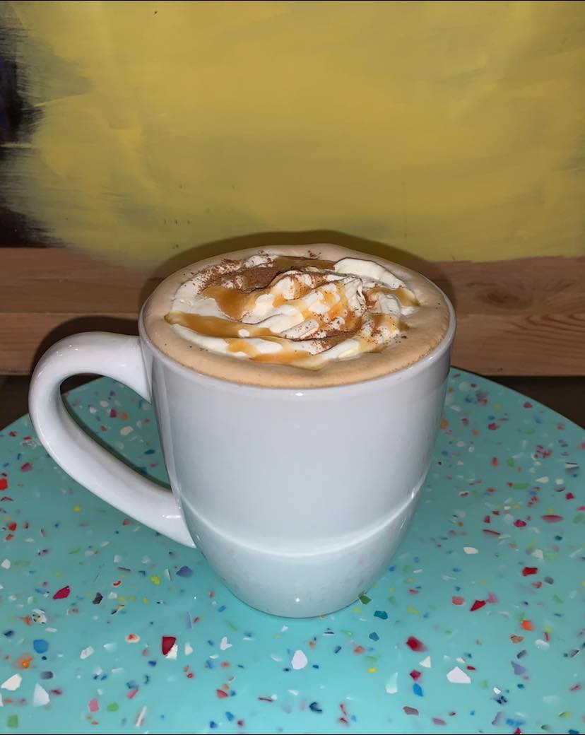 White mug with a caramel latte on a teal surface with a yellow backdrop.