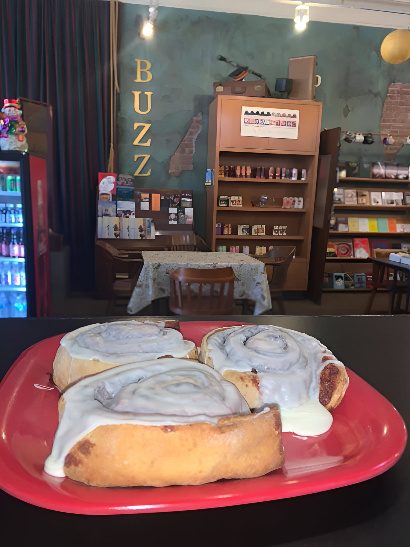 Three frosted cinnamon rolls on a red plate in a cafe. Behind, a store counter and shelves of books.