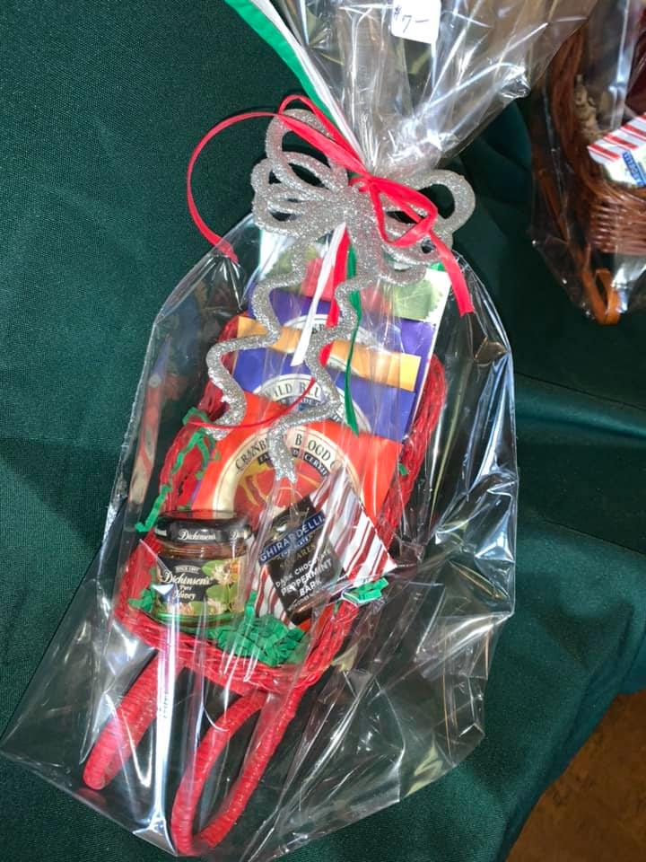 Wrapped gift basket with red sleigh-shaped ornament, clear wrapping, and silver and red ribbon against a green background.