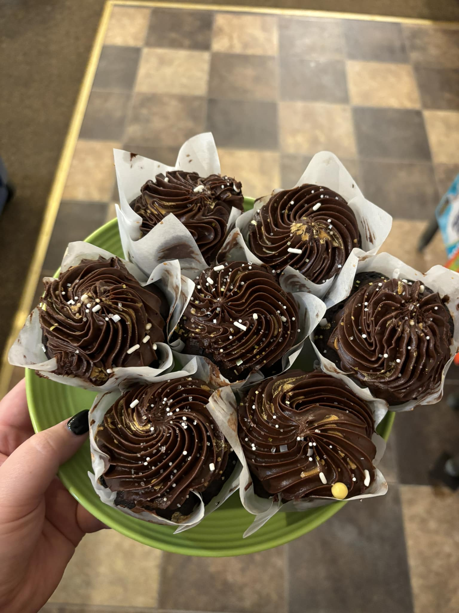 A hand holding a green plate with seven chocolate cupcakes in white paper liners, swirled frosting, and sprinkles.