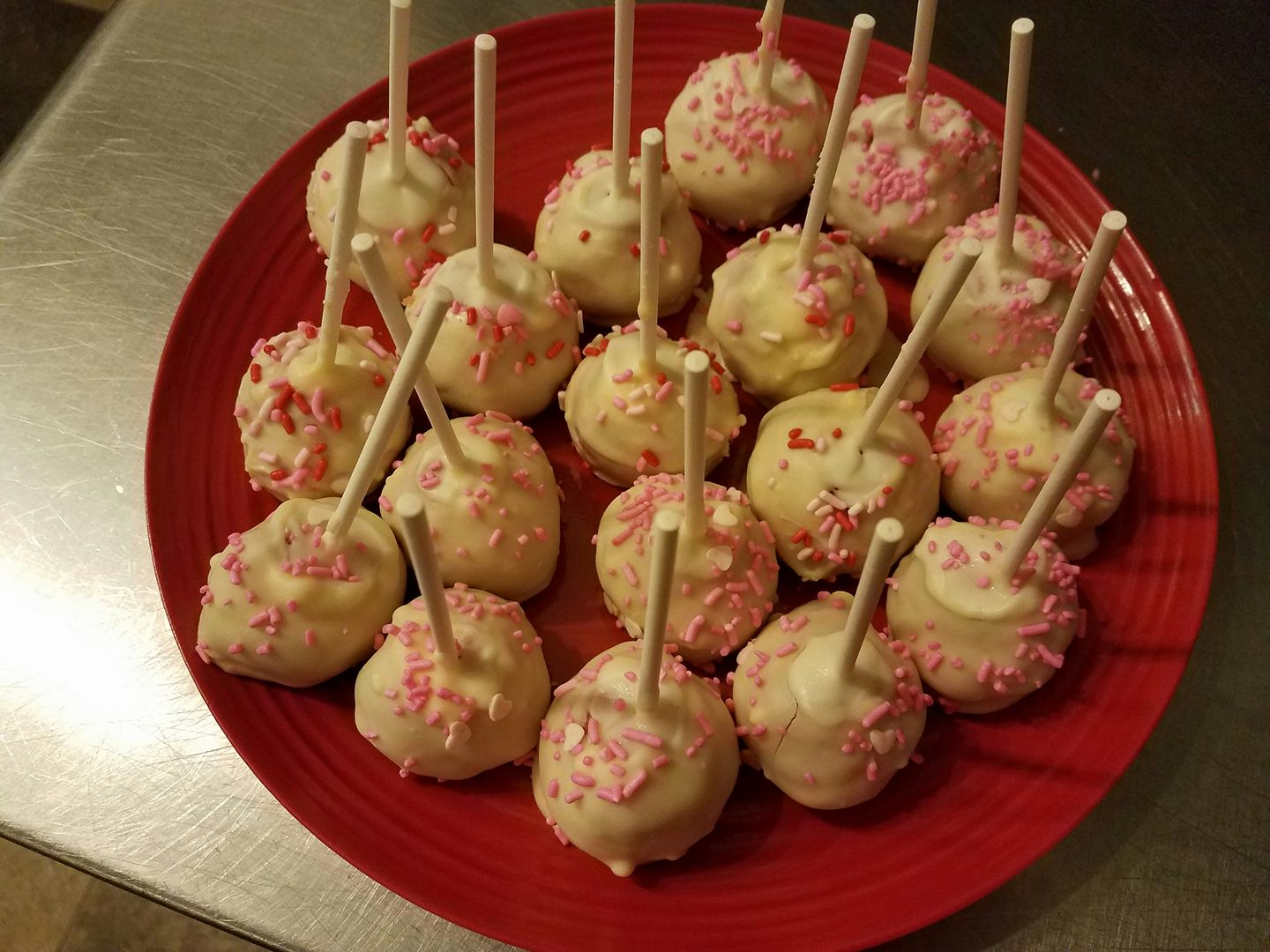 Cake pops on a red plate, coated in white frosting and pink sprinkles, with wooden sticks.