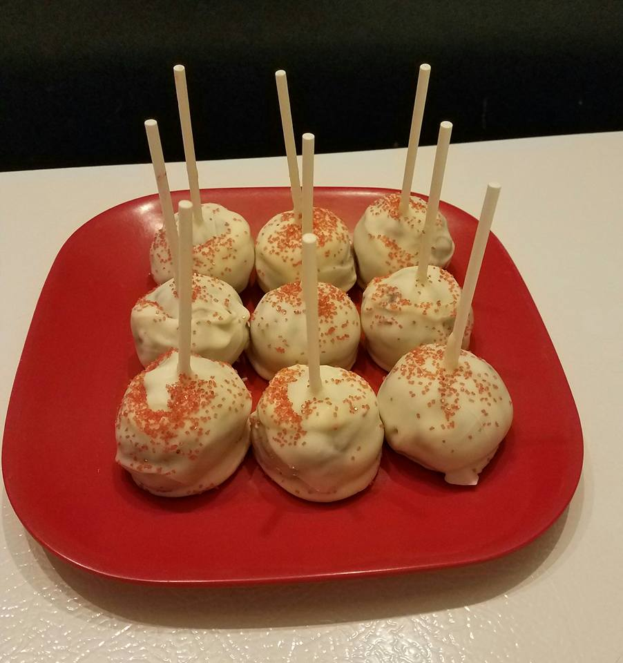 Nine white-frosted cake pops with red sprinkles on a red plate, with wooden sticks.