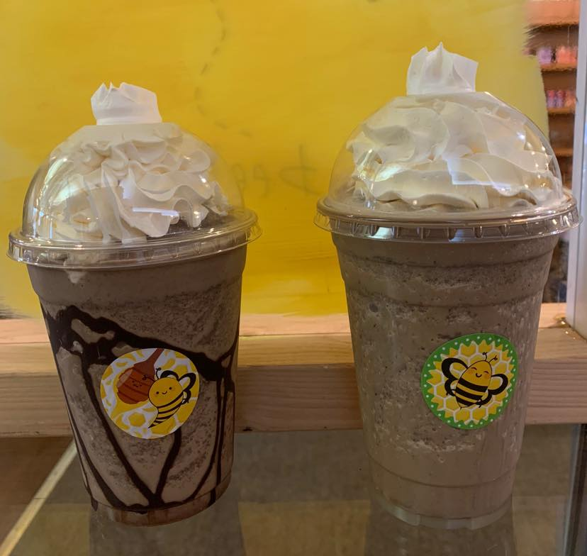 Two blended beverages with whipped cream topping, in clear plastic cups, set against a yellow background.