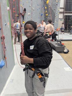 A smiling boy wearing a climbing harness stands beside an indoor climbing wall. with help from Beyond Boundaries
